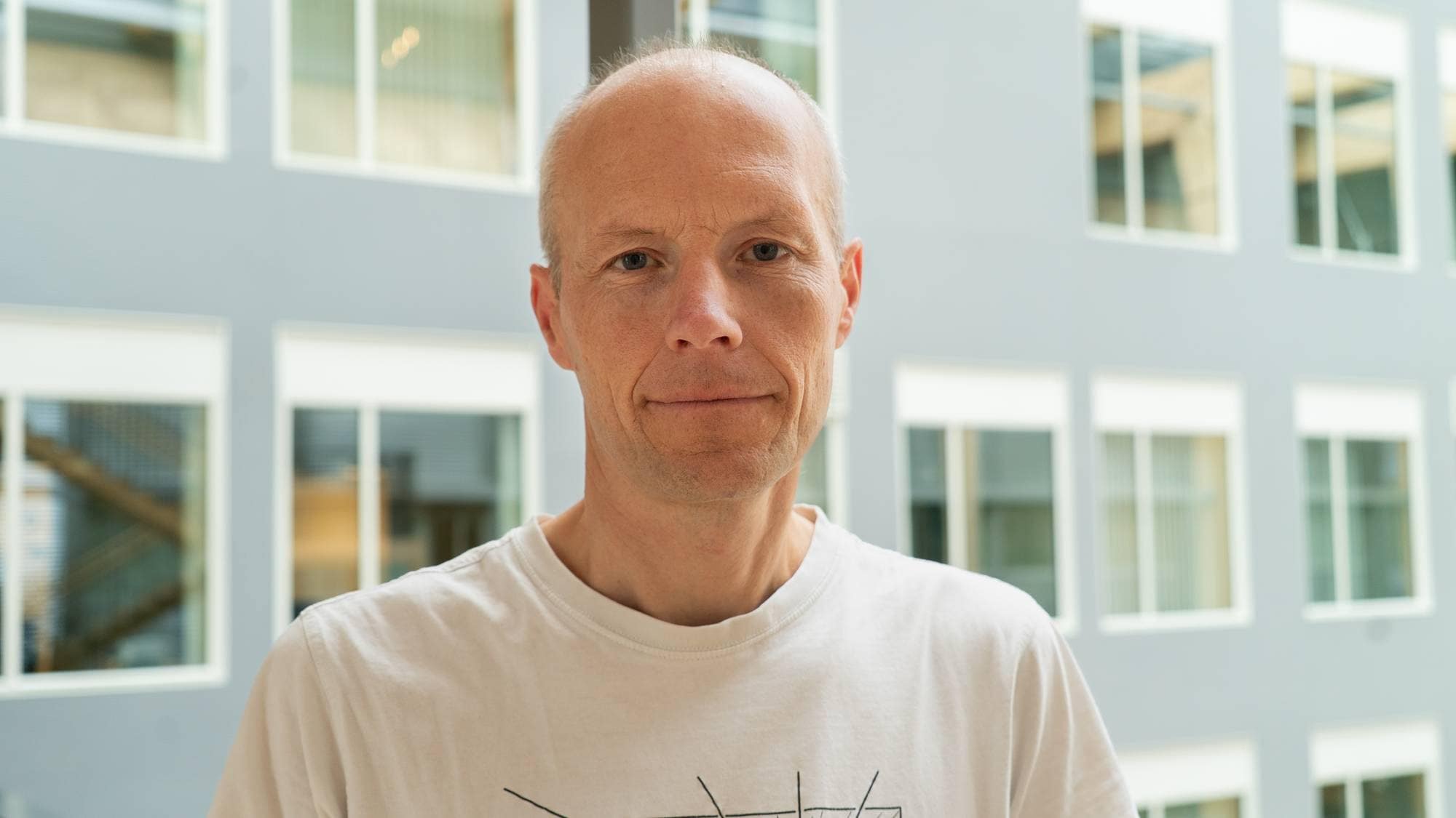 En mann i hvit t-skjorte står oppstilt i et portrett foran en foaje.