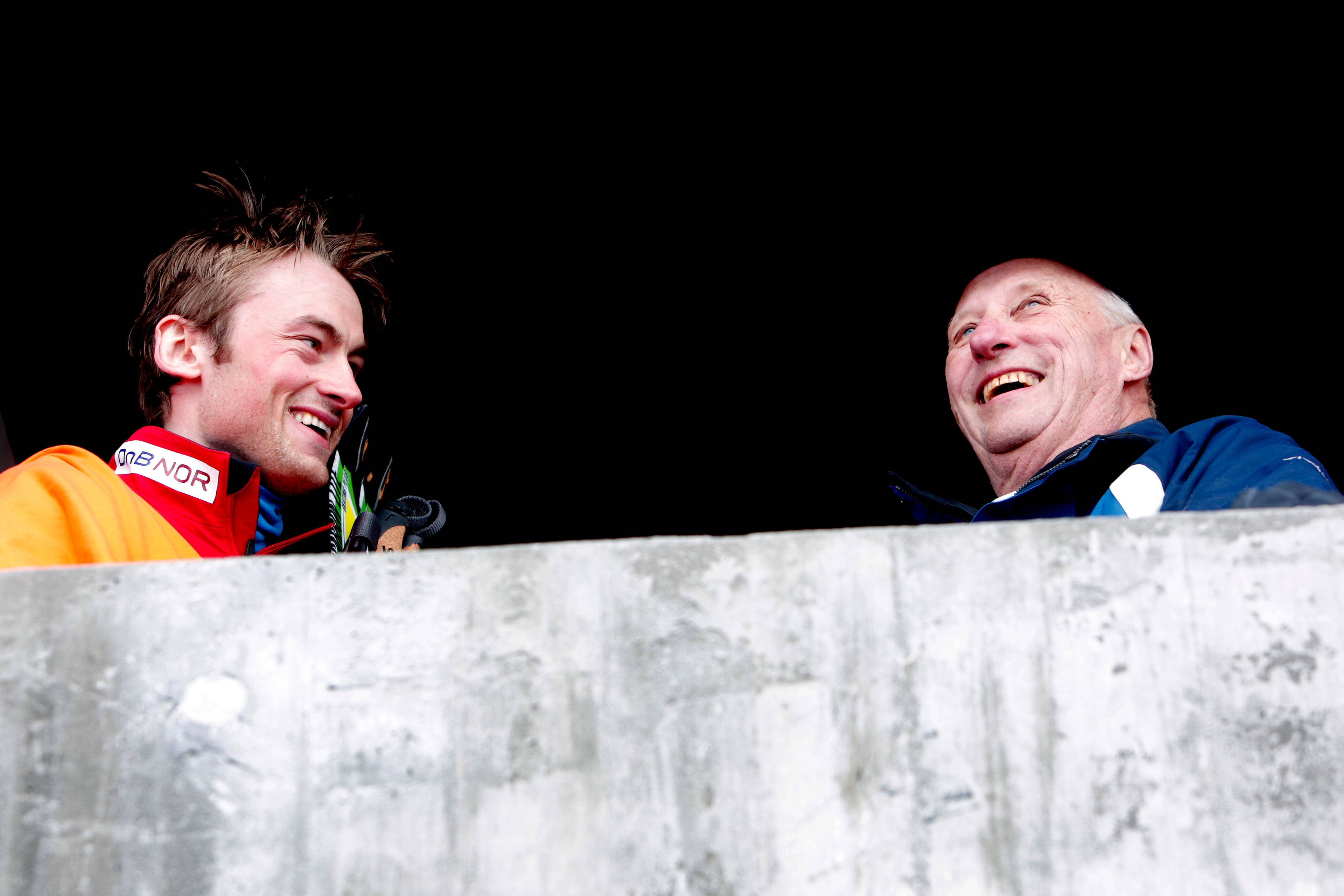 Petter Northug smiler til en eldre mann ved siden av seg bak en lav betongmur. (Bildebeskrivelsen er laget av en KI-tjeneste)