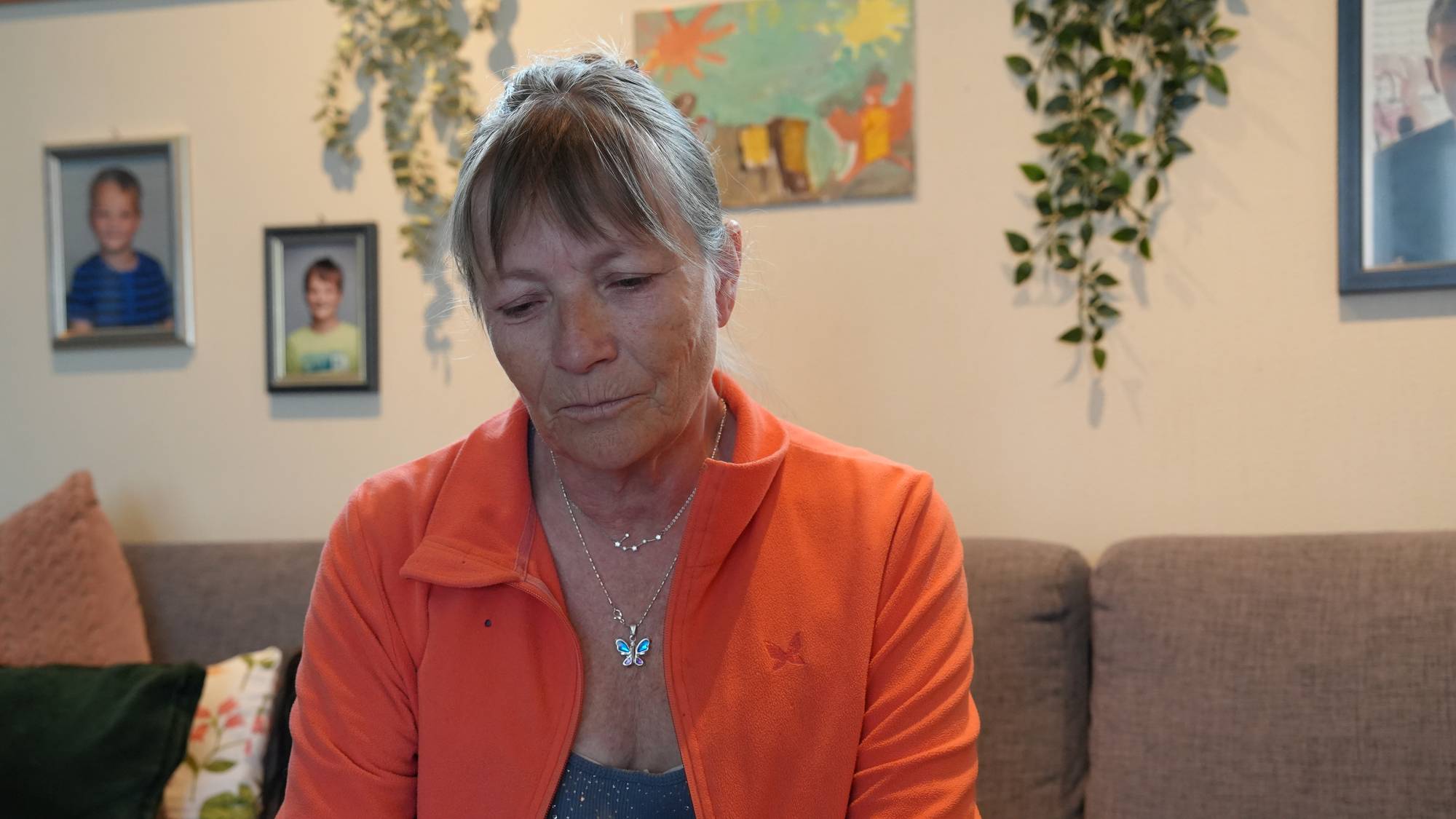 Kvinne i oransje jakke sitter i sofa, ser ned (i fotoalbum), bilder på veggen bak 