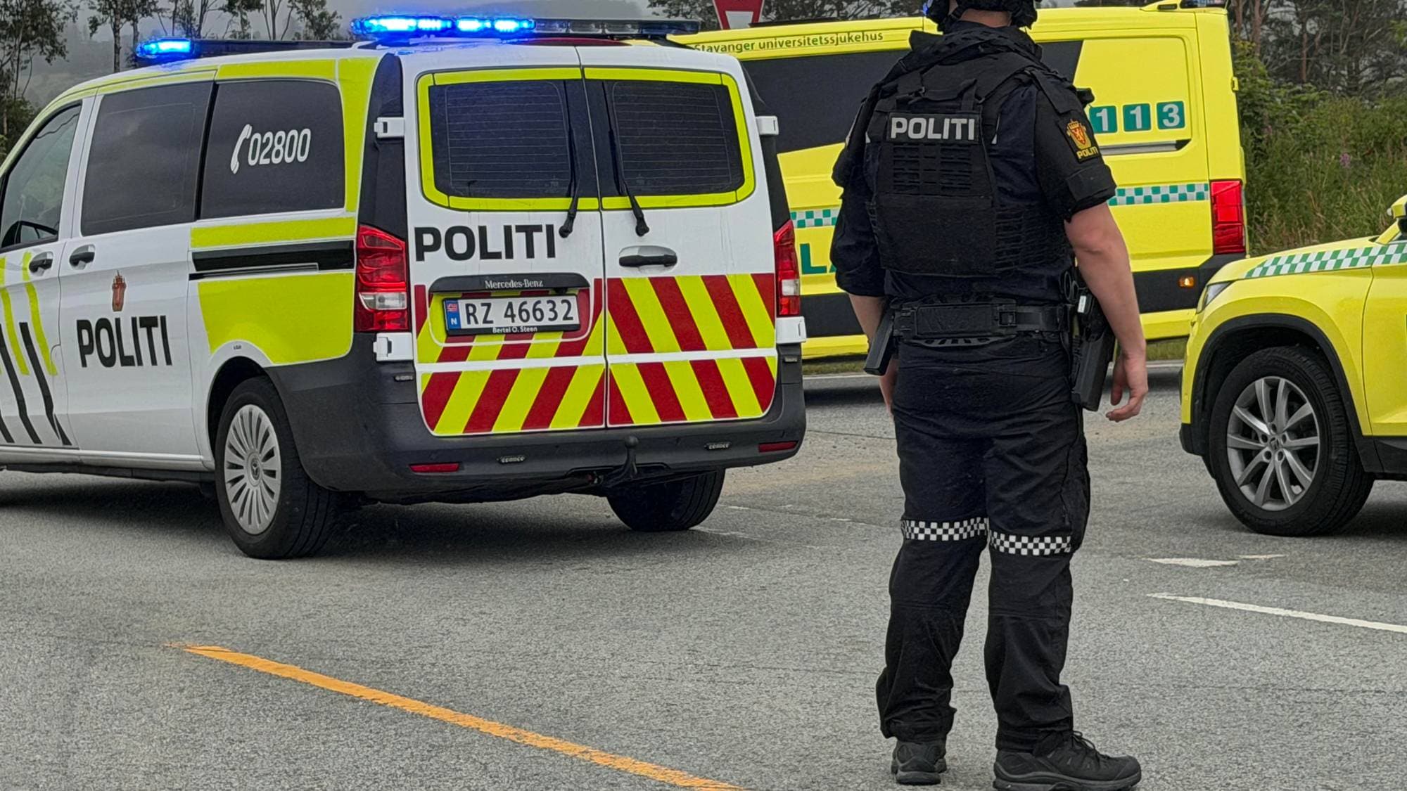 En politimann står med ryggen til. En politibil og to ambulanser står i bakgrunnen. 
