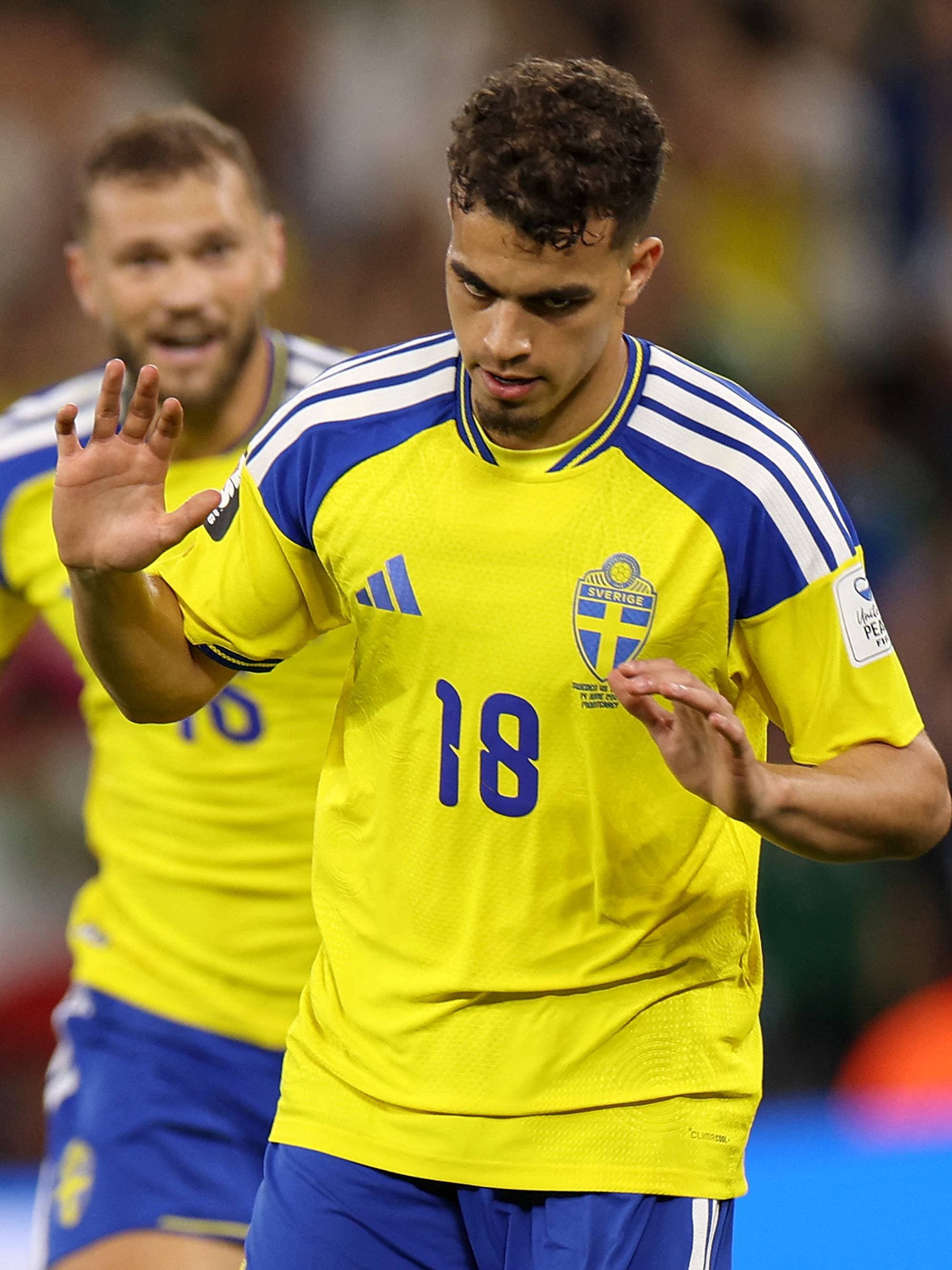 Two Swedish football players in yellow jerseys with blue trim stand on a field. The player in the foreground, wearing number 18, raises his hand. Behind him, another player in a similar jersey smiles. The background shows blurred spectators in a stadium. (Bildebeskrivelsen er laget av en KI-tjeneste)
