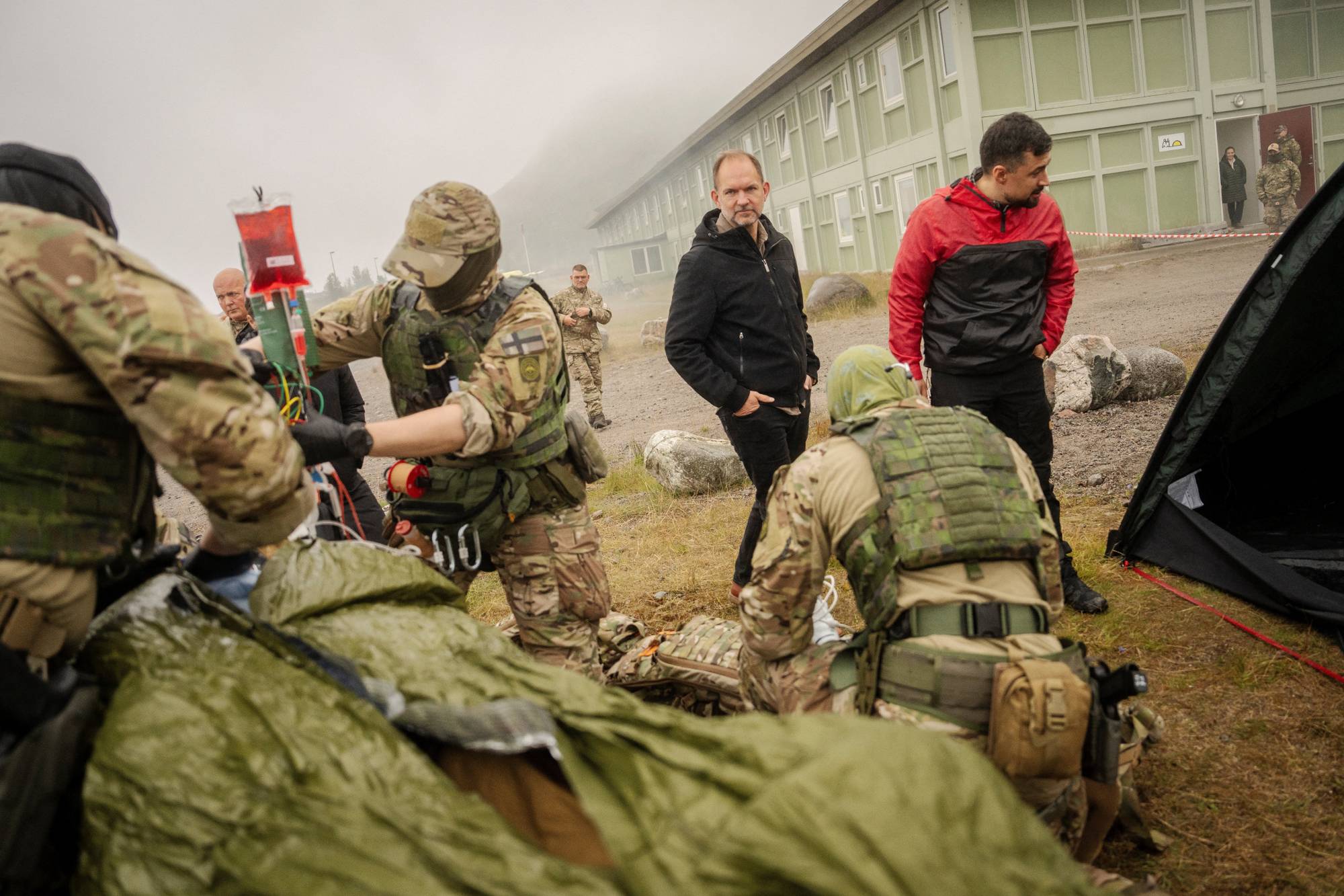 Soldater i kamuflasje gir medisinsk behandling til en person på en båre utendørs ved en militærleir. (Bildebeskrivelsen er laget av en KI-tjeneste)
