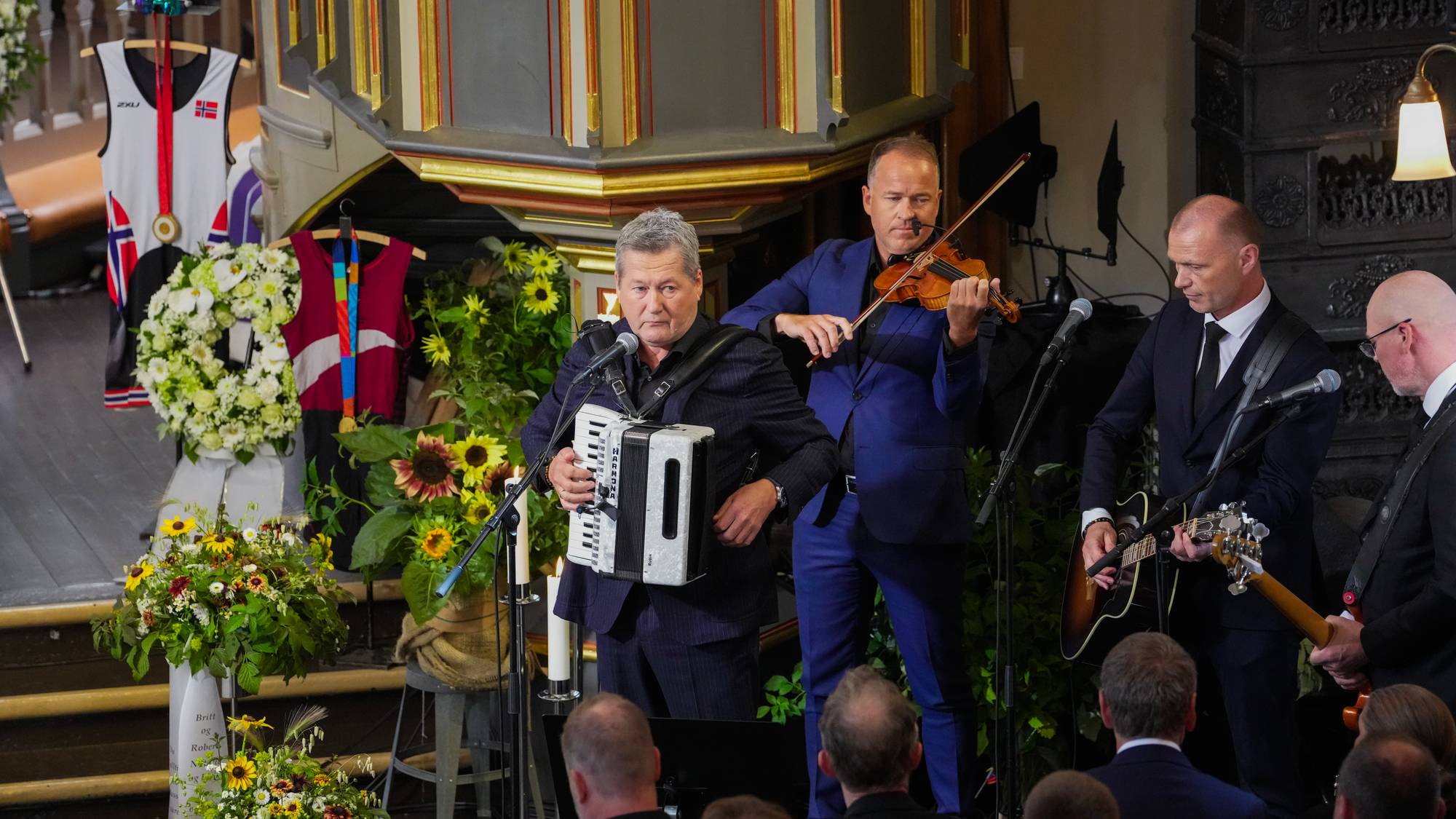 Musikere spiller trekkspill, fele og gitar under en seremoni i en kirke med blomsterdekorasjoner.