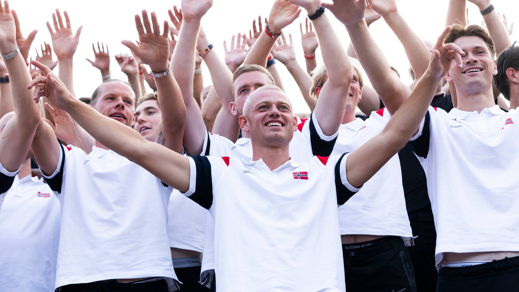 En gruppe idrettsutøvere i hvite drakter med det norske flagget står med armene løftet i været. (Bildebeskrivelsen er laget av en KI-tjeneste)