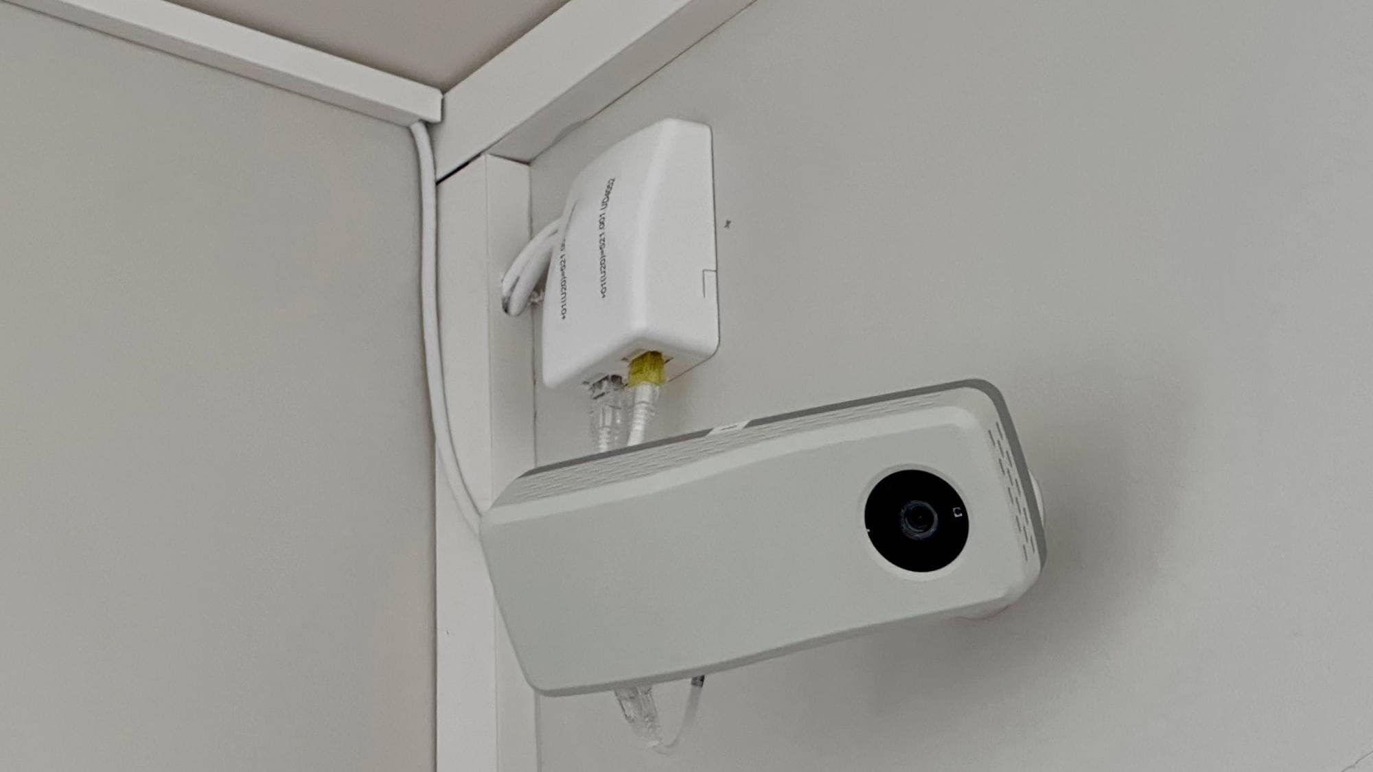 Overvåkningskamera montert i et hjørne på en hvit vegg, koblet til en veggkontakt med kabler. (Bildebeskrivelsen er laget av en KI-tjeneste)