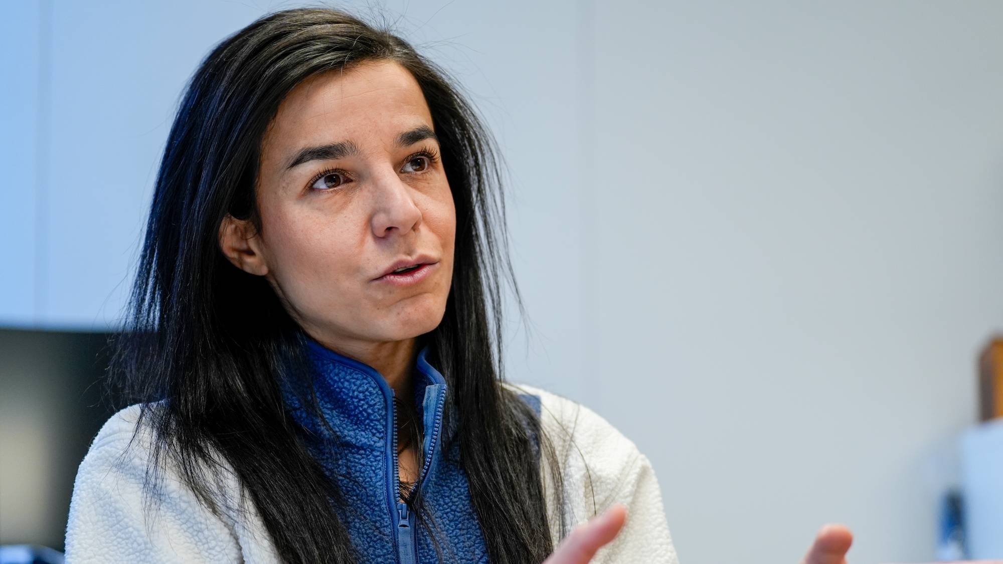En person med langt, svart hår sitter i et rom og gestikulerer med hendene. De har på seg en blå fleecejakke og åpner opp om et tema. Bak dem kan en datamaskin og en innredet vegg sees. Området virker pent og profesjonelt. (Bildebeskrivelsen er laget av en KI-tjeneste)