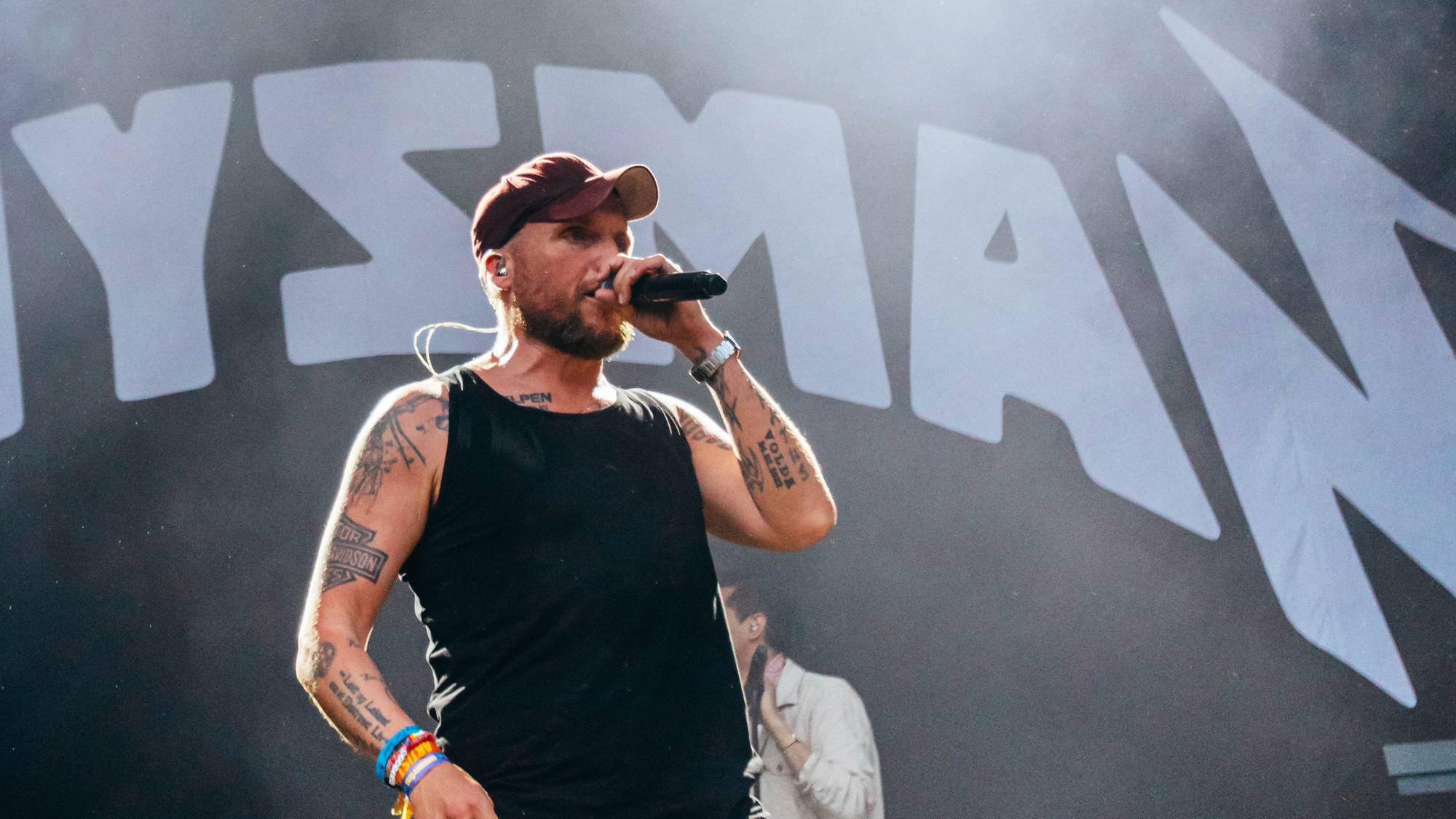 En mann med keps, tetoveringer og blå dongerishorts står på en scene og holder en mikrofon. Bak ham er det en stor banner med tekst. Det er røyk i luften og lyskastere på scenen. (Bildebeskrivelsen er laget av en KI-tjeneste)