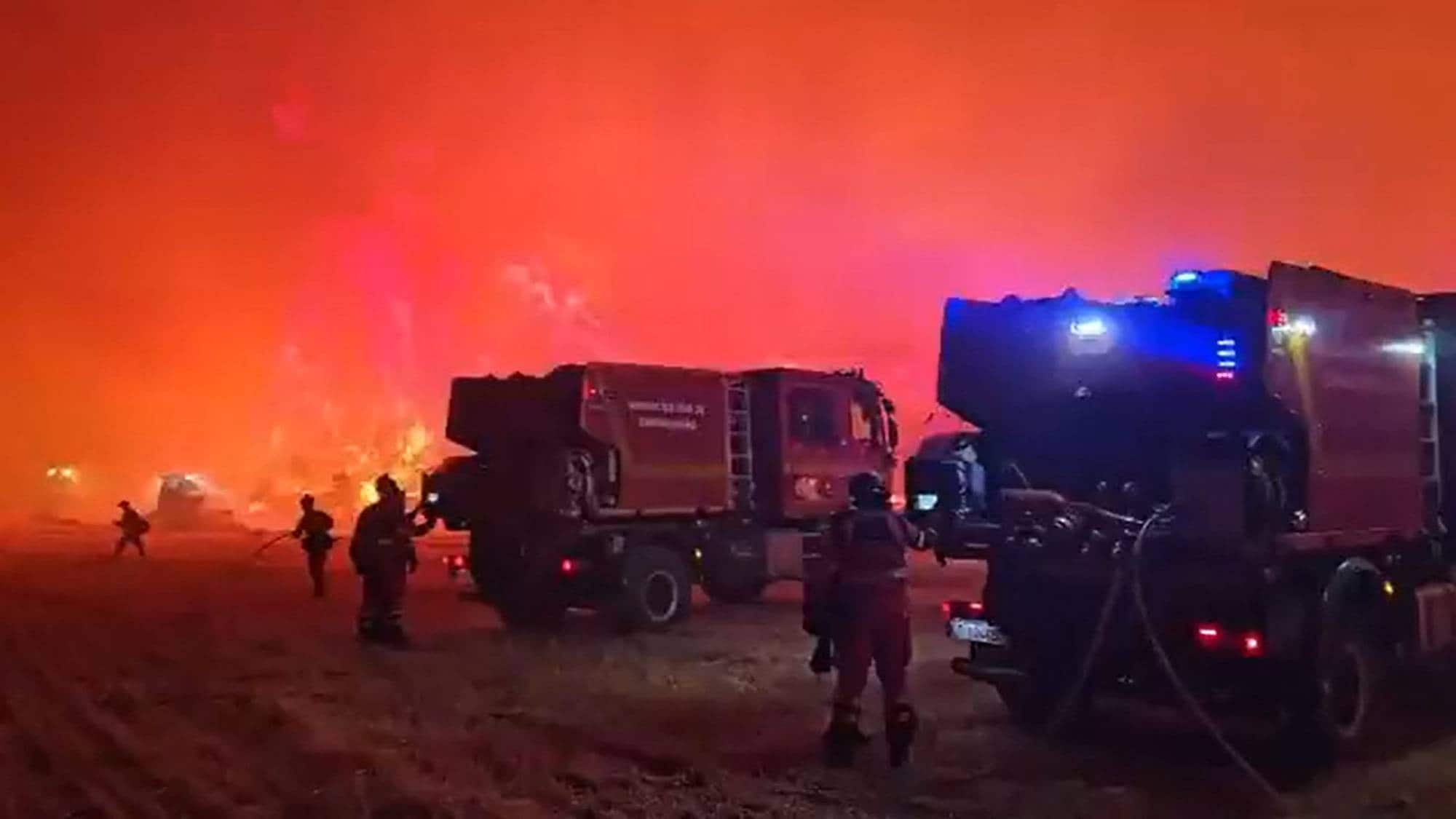 Brannmannskaper arbeider ved store brannbiler foran en kraftig flammevegg om natten. (Bildebeskrivelsen er laget av en KI-tjeneste)