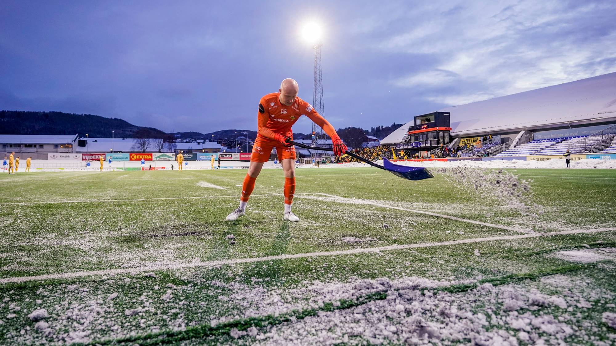 Målvakt fjerner snø på kunstgressbane. Spilleren bruker en spade til å skuffe bort snø som har lagt seg på banen. Stadionet er dekket av snø. Flere spillere i gult og oransje er synlige i bakgrunnen. (Bildebeskrivelsen er laget av en KI-tjeneste)