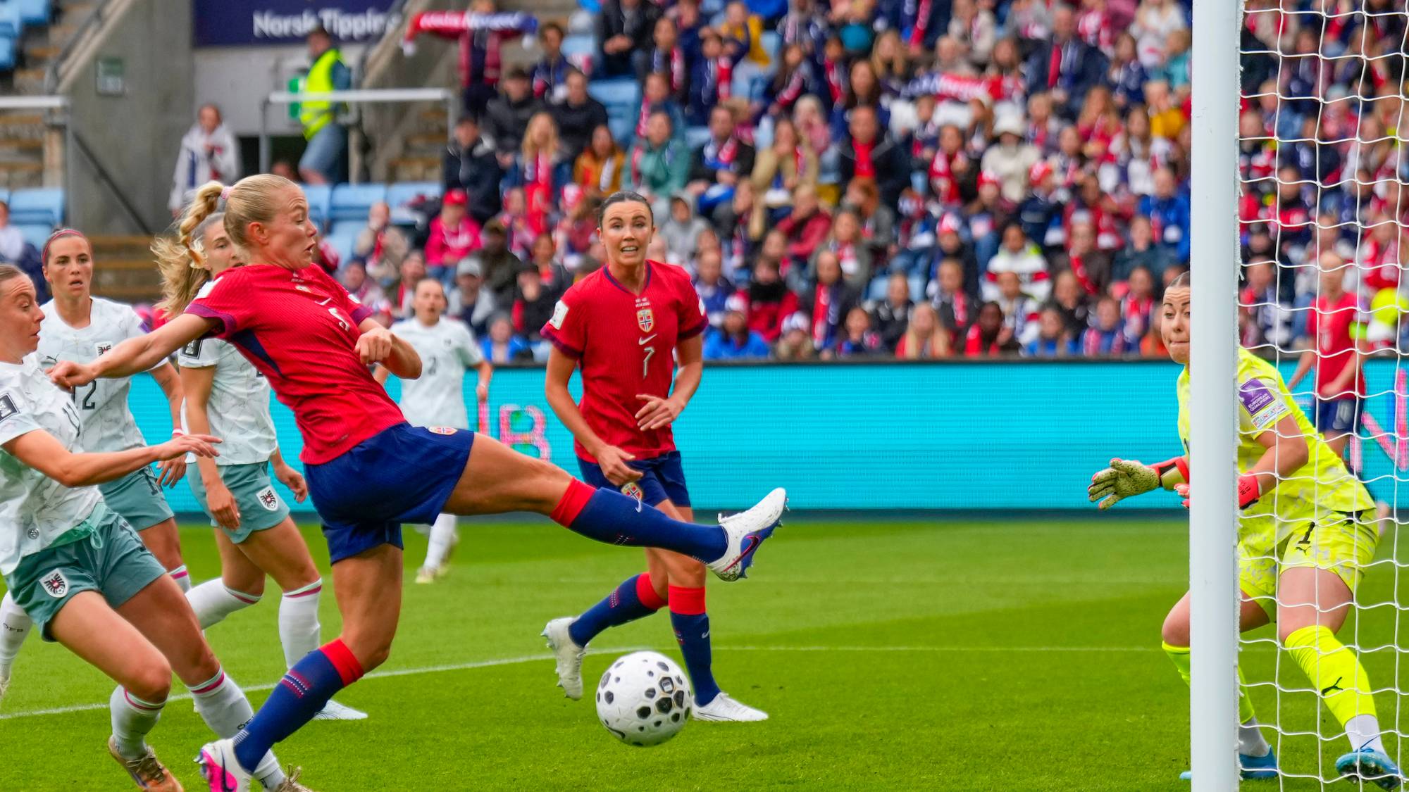 En fotballspiller i rødt sparker til ballen. En keeper i gult er klar ved målnettet. Andre spillere i hvitt og blått er i bakgrunnen. Publikum sitter på tribunen. (Bildebeskrivelsen er laget av en KI-tjeneste)