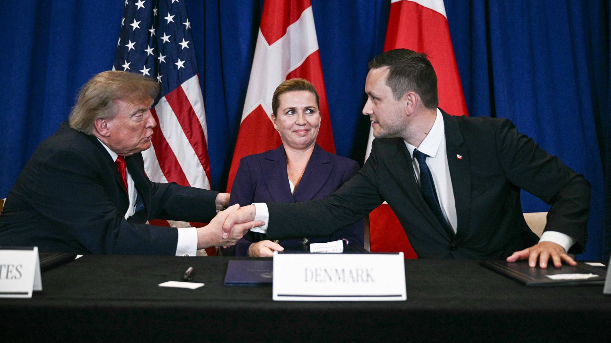 Donald Trump og en dansk representant håndhilser foran Mette Frederiksen med flagg i bakgrunnen. 