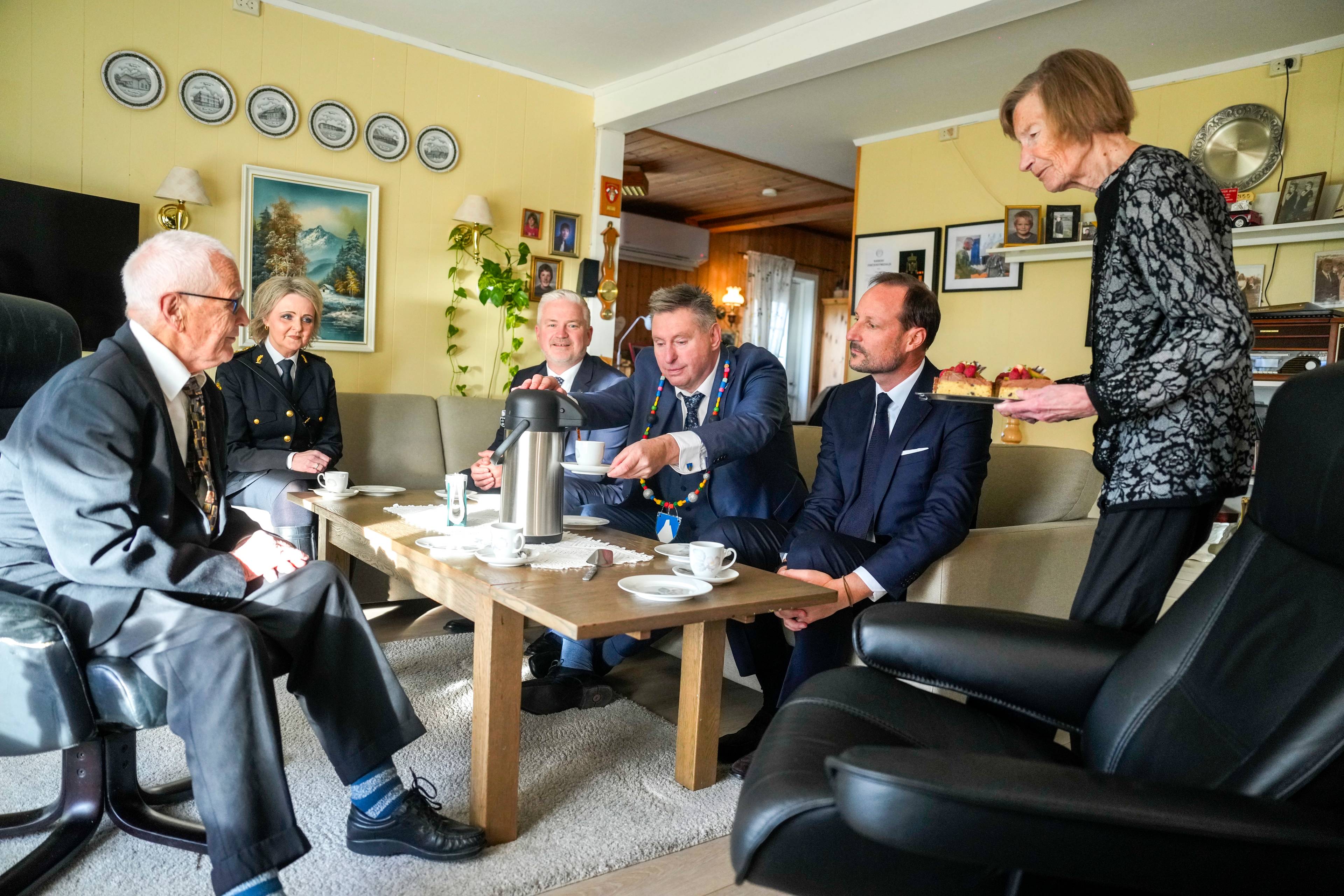 Kronprins Haakon og ordfører sitter sammen med Magne Øymo i en stue mens de blir servert kaffe. (Bildebeskrivelsen er laget av en KI-tjeneste)