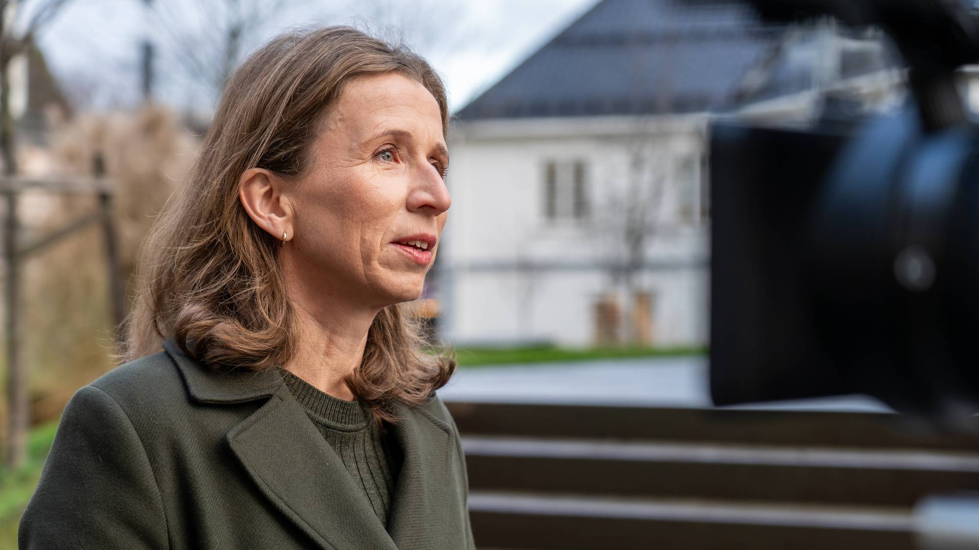 En kvinne står foran en kamerautstyr i en utendørs setting. Hun har på seg en mørkegrønn jakke og ser mot kameraet. I bakgrunnen kan man se en bygning og trær, og bakken er støpt med trapper. Det er klart vær, og omgivelsene gir en uformell kontekst til situasjonen. (Bildebeskrivelsen er laget av en KI-tjeneste)