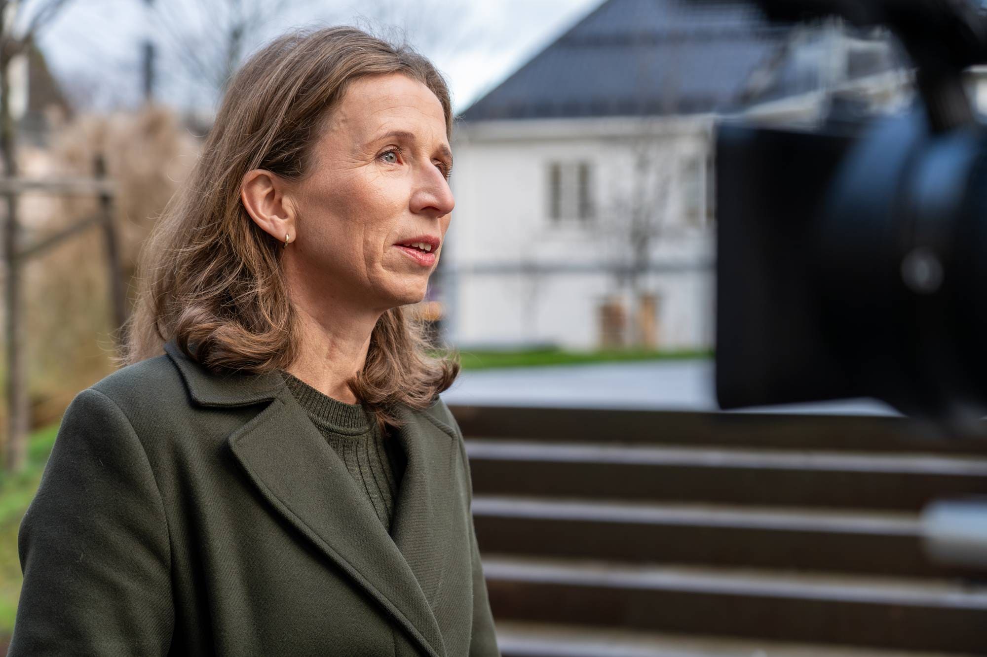 En kvinne står foran en kamerautstyr i en utendørs setting. Hun har på seg en mørkegrønn jakke og ser mot kameraet. I bakgrunnen kan man se en bygning og trær, og bakken er støpt med trapper. Det er klart vær, og omgivelsene gir en uformell kontekst til situasjonen. (Bildebeskrivelsen er laget av en KI-tjeneste)