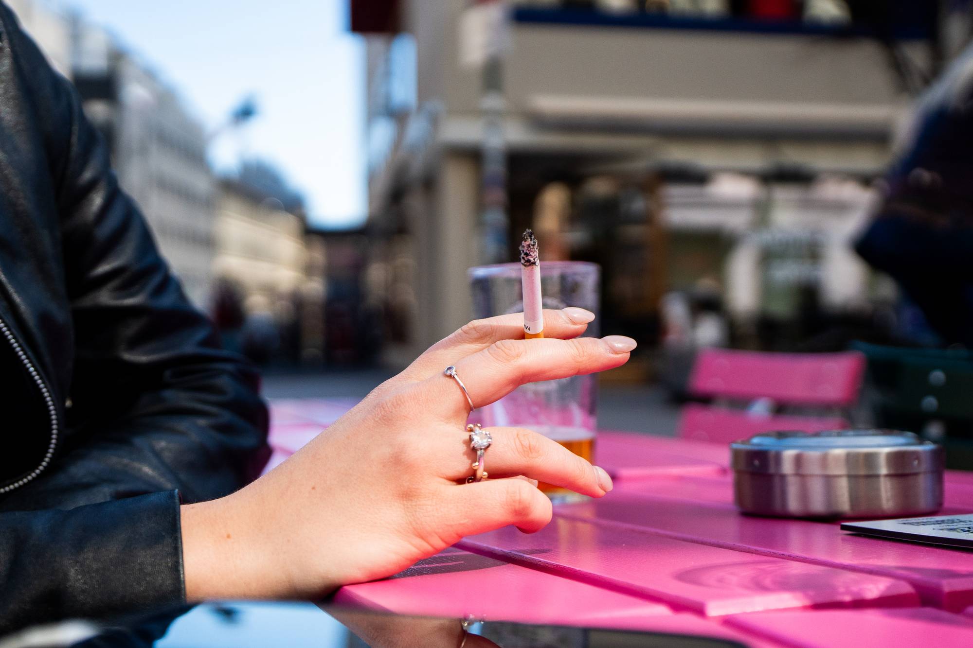 En kvinne holder en sigarett som brenner. Hun sitter ved et rosa bord på en uteservering. En glass med øl står på bordet. I bakgrunnen sees en bygate med bygninger. (Bildebeskrivelsen er laget av en KI-tjeneste)
