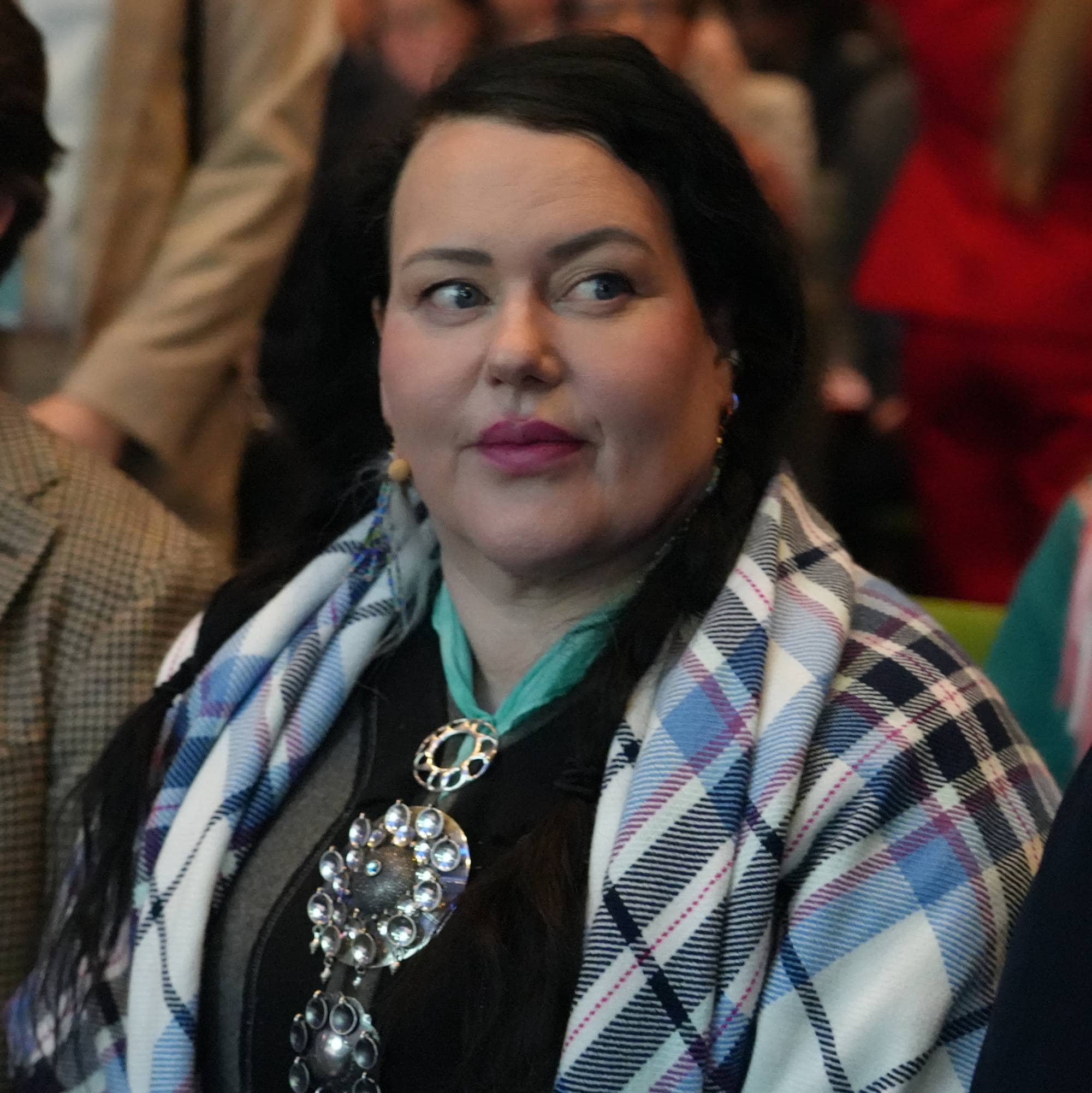Bildet viser en gruppe mennesker som sitter i et auditorium. Flere personer er synlige, med en kvinne i front som har på seg en fargerikt sjal og sølje. I bakgrunnen er det en kvinne som står med et lyst antrekk. Det er en uformell atmosfære med mennesker som ser ut til å delta på et arrangement. (Bildebeskrivelsen er laget av en KI-tjeneste)