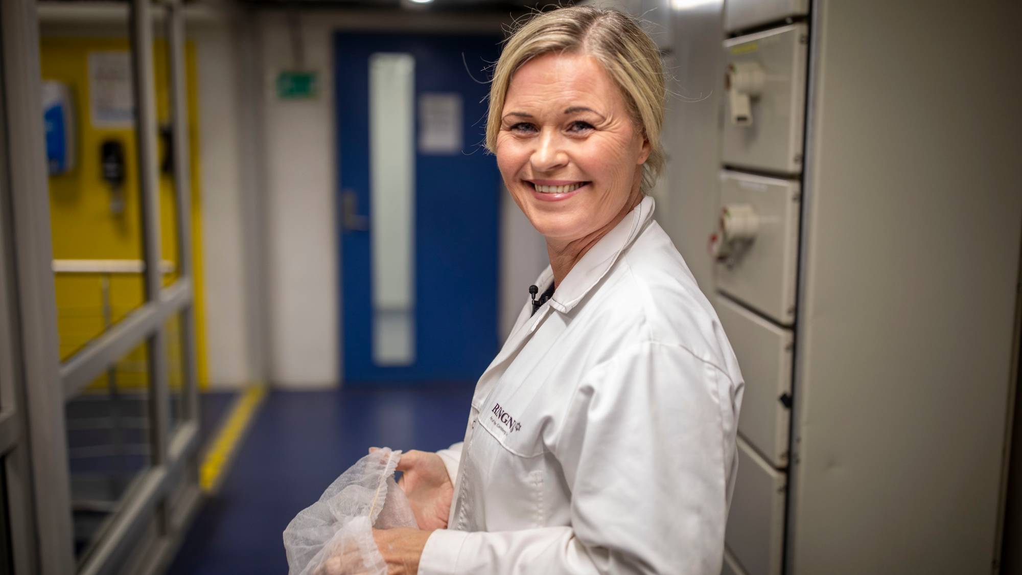 Smilende kvinne med lyst hår og hvit frakk foran en blå dør.