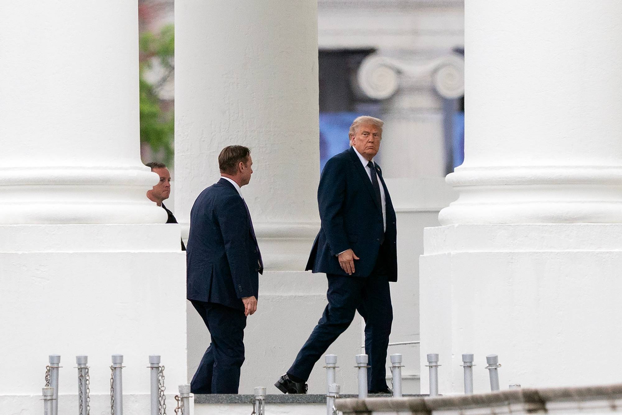 Donald Trump mellom store, hvite søyler. Bak ham to menn som ser ut som sikkerhetsvakter.