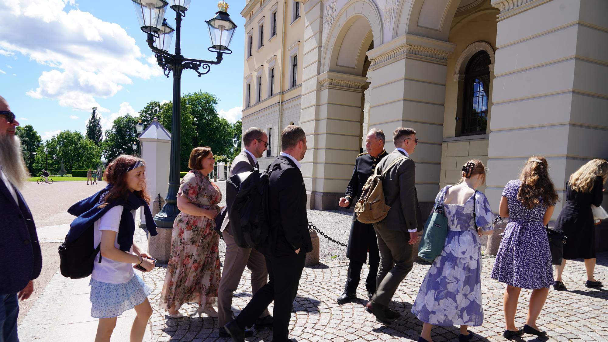 En gruppe mennesker, inkludert en ung jente med ryggsekk, går over en brosteinsbelagt gårdsplass. De nærmer seg en stor, lys bygning med buede innganger. En mann i uniform går blant de andre. (Bildebeskrivelsen er laget av en KI-tjeneste)