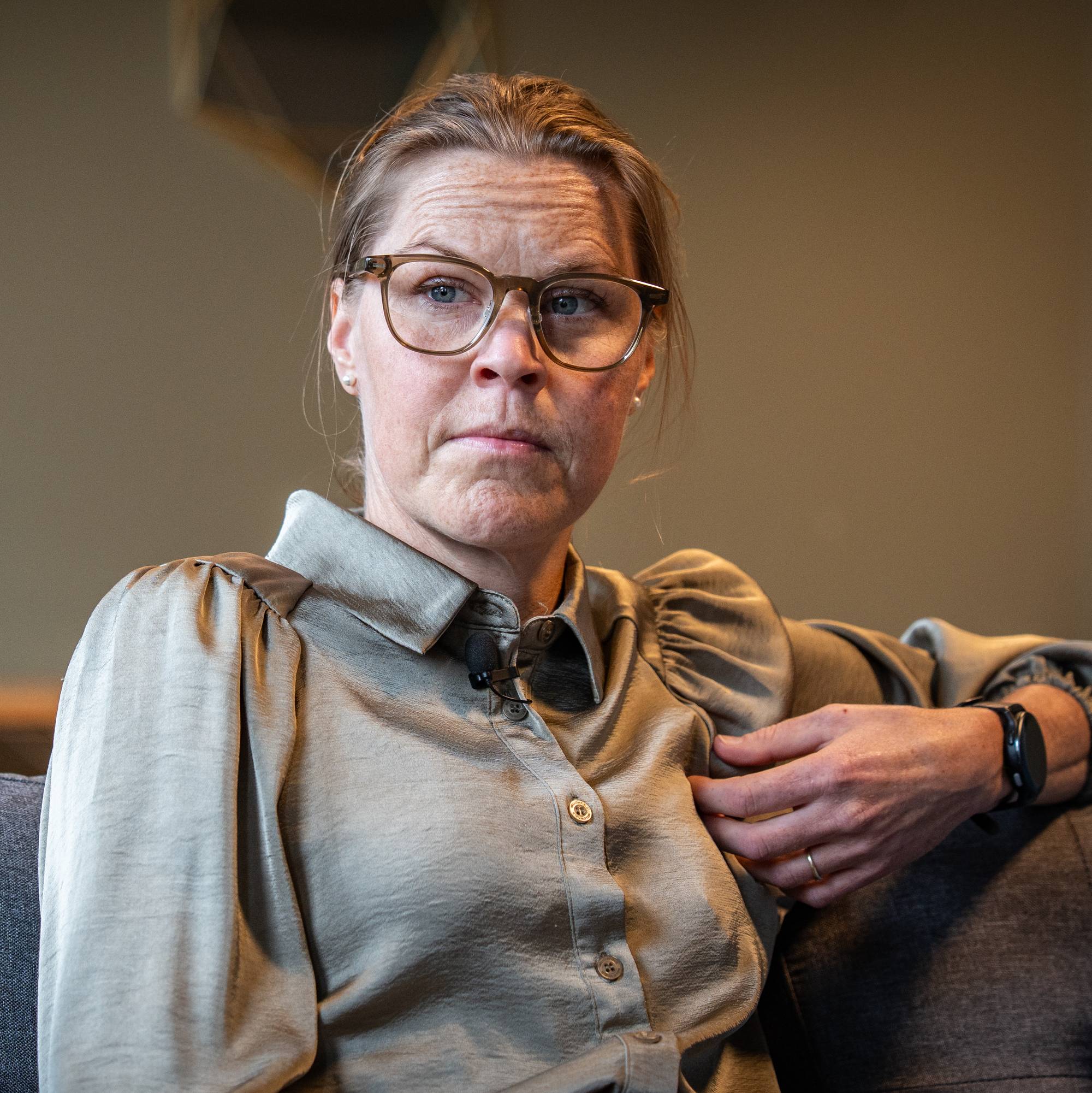 En kvinne med briller og grønn bluse sitter i en sofa. I bakgrunnen ser man en vegg. 