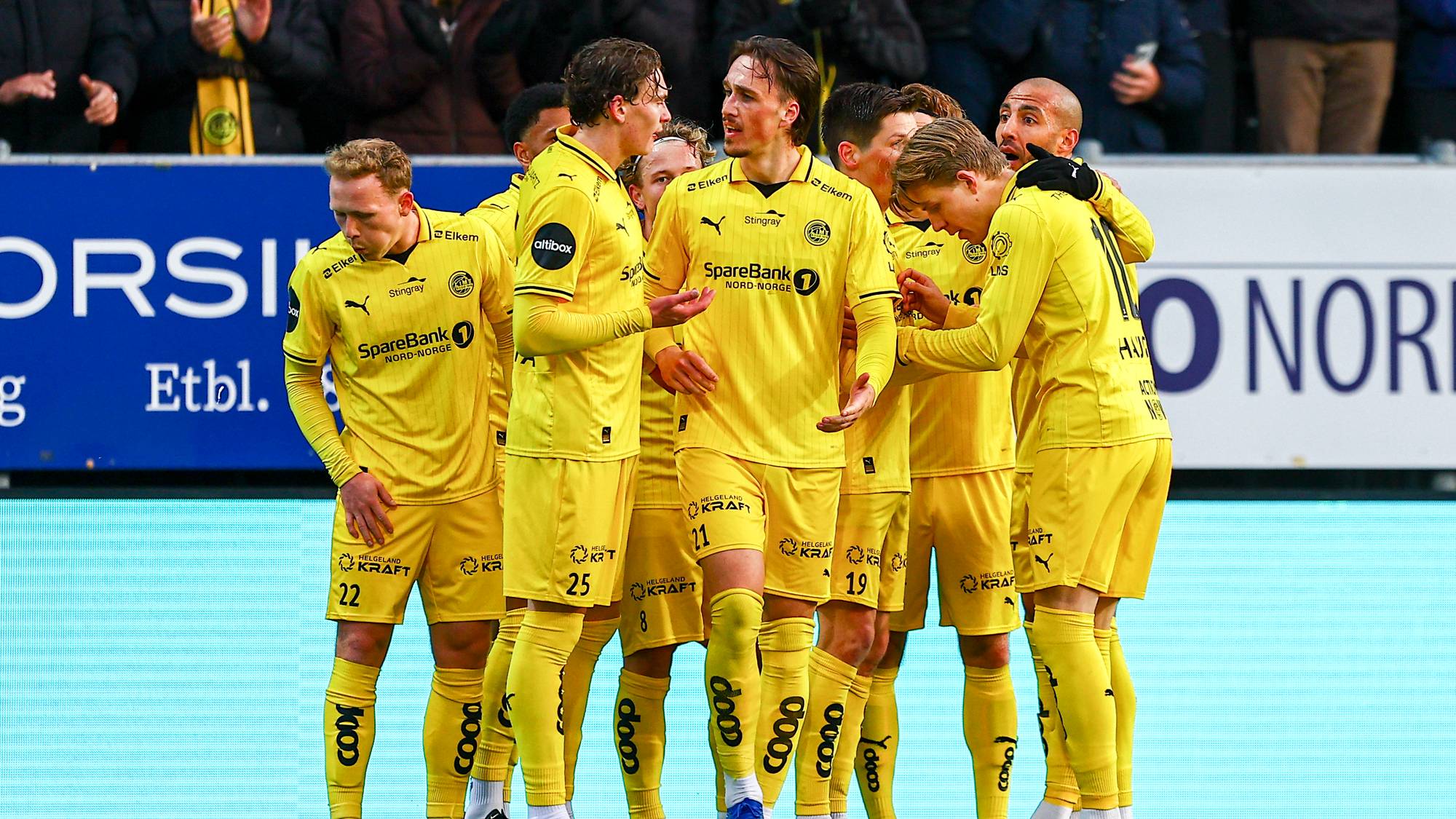 Fotballspillere i gule drakter står samlet på en bane. Spillerne er omkranset av medspillere og ser ut til å diskutere. Bak dem er det tribuner med publikum. Bakgrunnen består av blå og lyseblå reklameskilt. (Bildebeskrivelsen er laget av en KI-tjeneste)