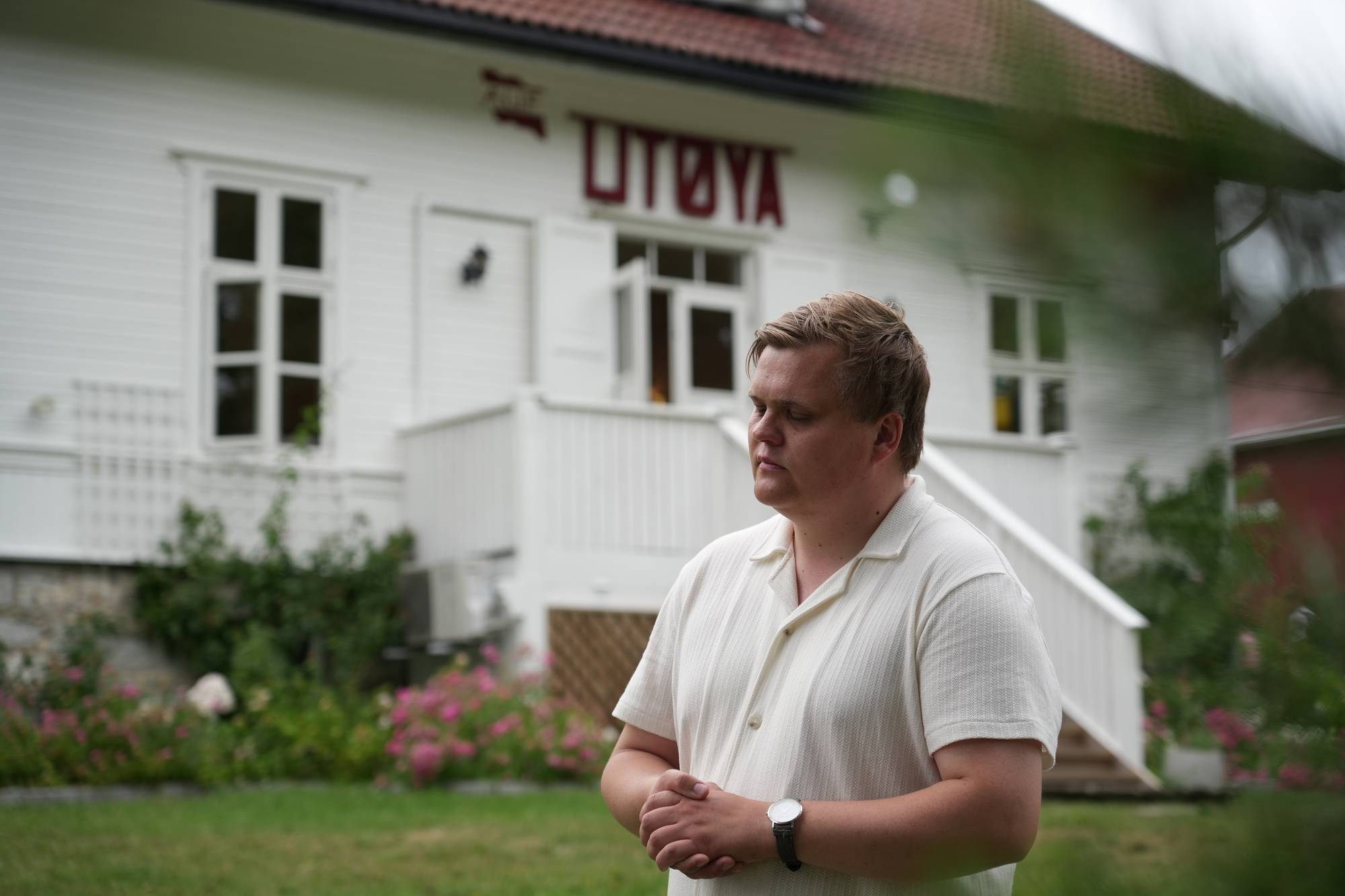 En mann med armene foldet foran seg står utenfor en hvit bygning med røde bokstaver som leser "UTØYA". Mannen har lukket øynene og ser ut til å være i ettertanke. En hage med blomster er synlig i forgrunnen. Bygningen er en hvit trebygning med flere vinduer og en trapp. (Bildebeskrivelsen er laget av en KI-tjeneste)