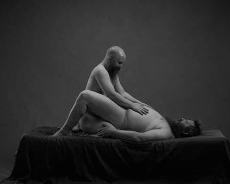 To nakne menn med langt skjegg ligger i senga. Den underste har bekkenet løftet og ligger med bena på hver side av den andre som står på kne.