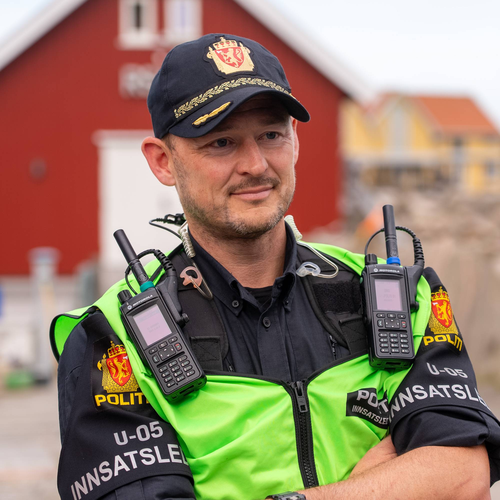 En pilitimann står med uniform med armene i kryss og med refleksvest. Det er et rødt hus i bakgrunnen. 