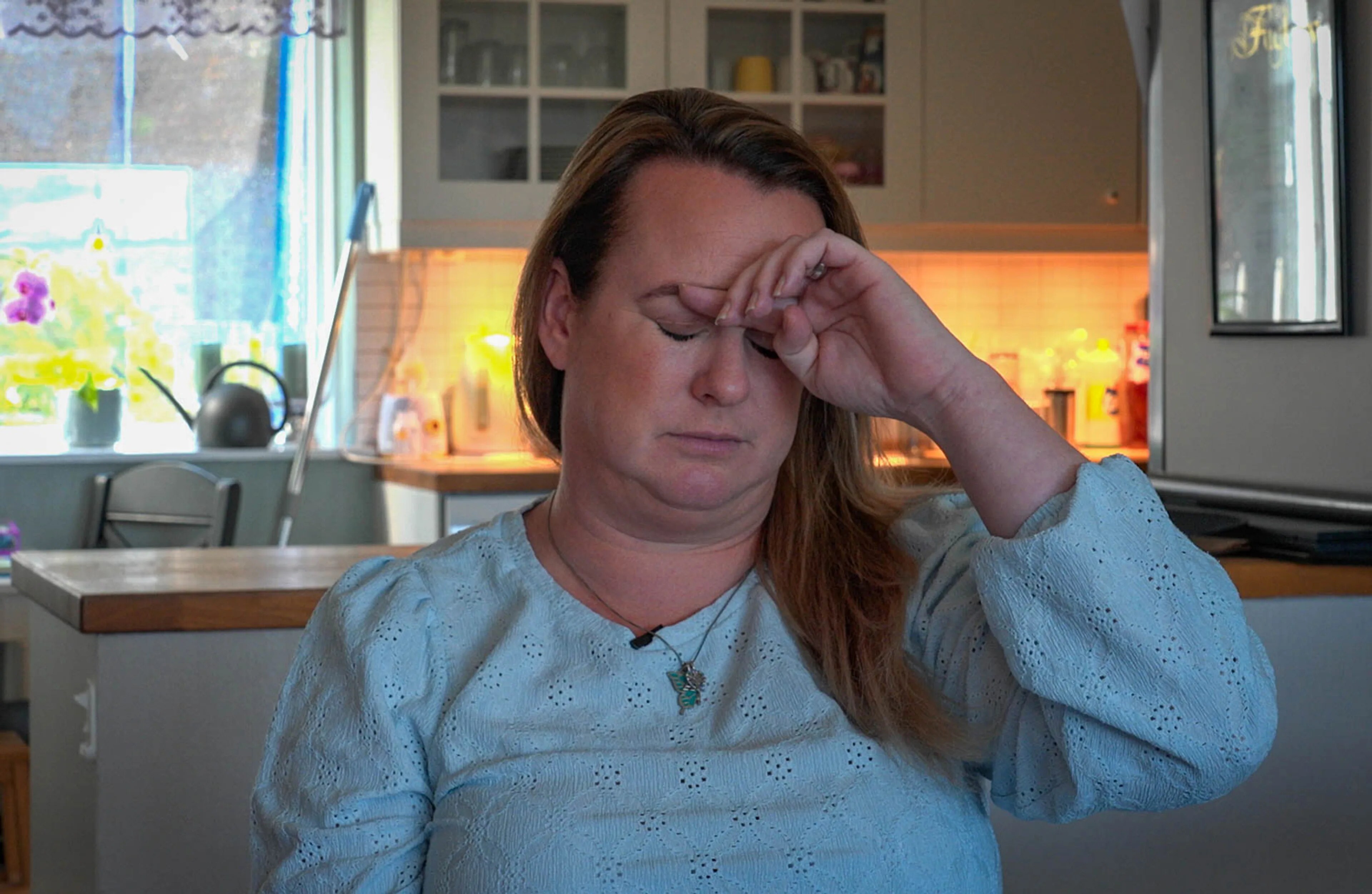 En kvinne med langt hår tar seg til pannen og ser lei seg ut. I bakgrunnen er det et hvitt kjøkken-