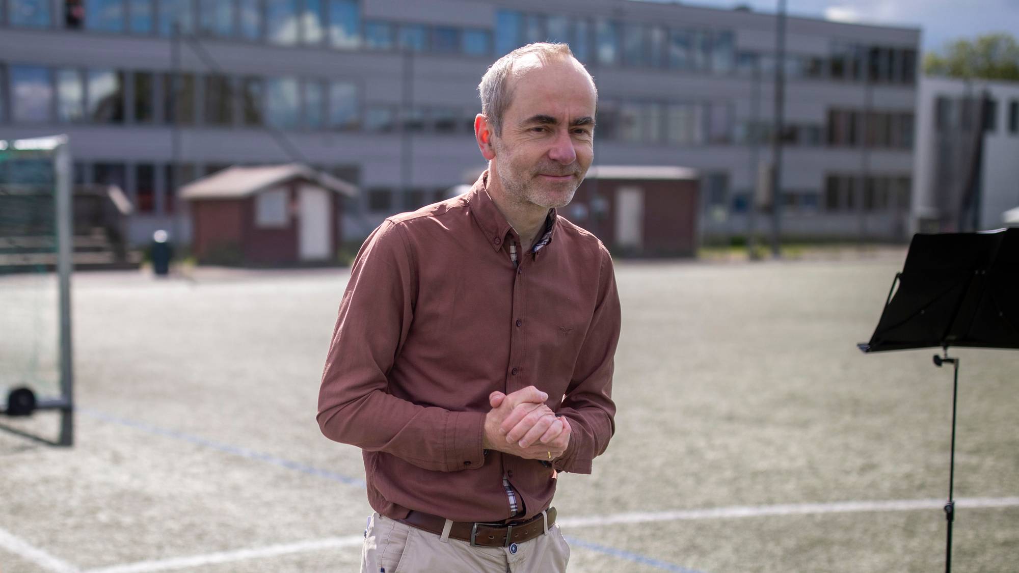 En mann i brun skjorte og lyse bukser står på en grønn kunstgressbane. Han har hendene knyttet foran seg. Til høyre står et note stativ, og til venstre skimtes en person i blått. I bakgrunnen er det en bygning og to små røde boder. (Bildebeskrivelsen er laget av en KI-tjeneste)