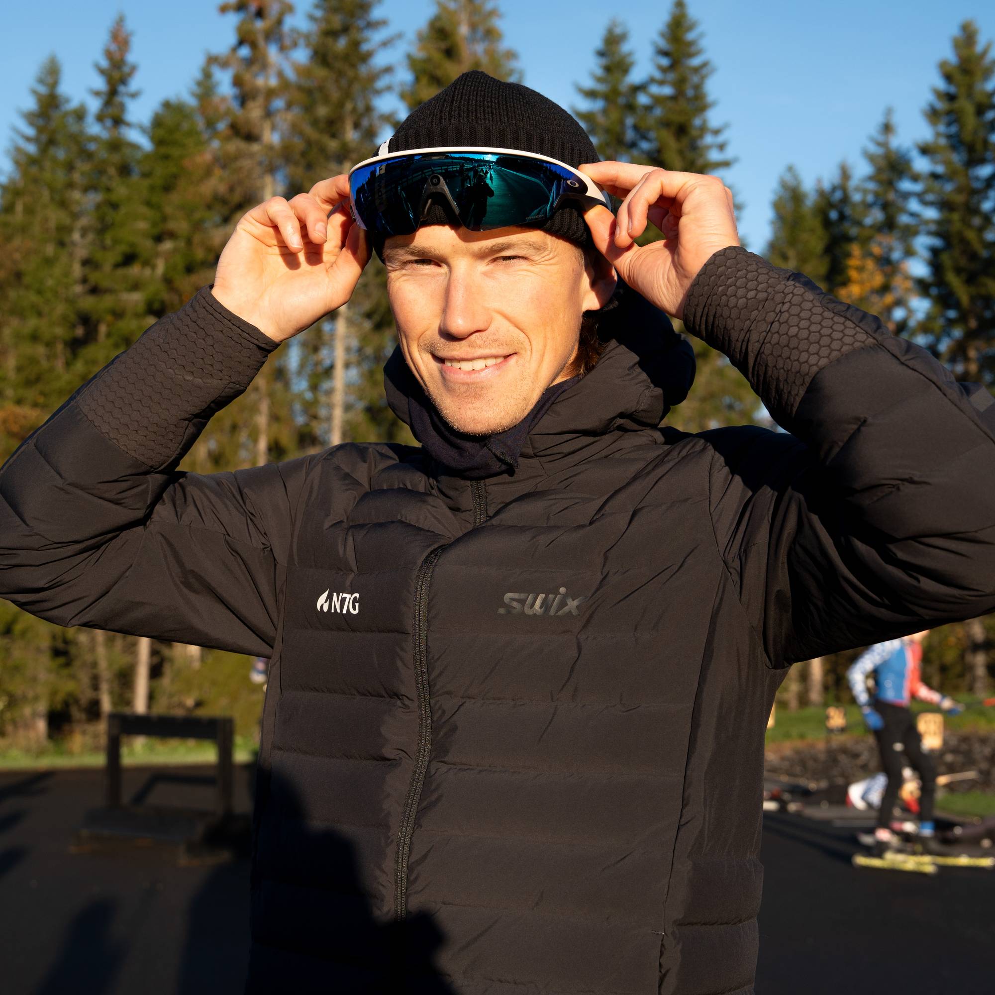 En smilende mann tar på seg sportsbriller ved en skiskytterarena med skog i bakgrunnen. Det er Meta-briller. (Bildebeskrivelsen er laget av en KI-tjeneste)