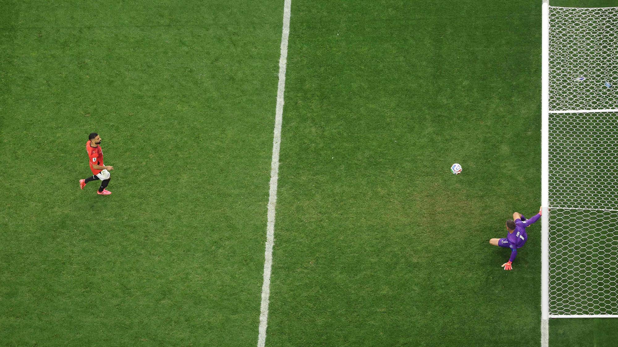 Fotballspiller i rød drakt skyter ballen mot mål, der keeperen i lilla drakt kaster seg. (Bildebeskrivelsen er laget av en KI-tjeneste)