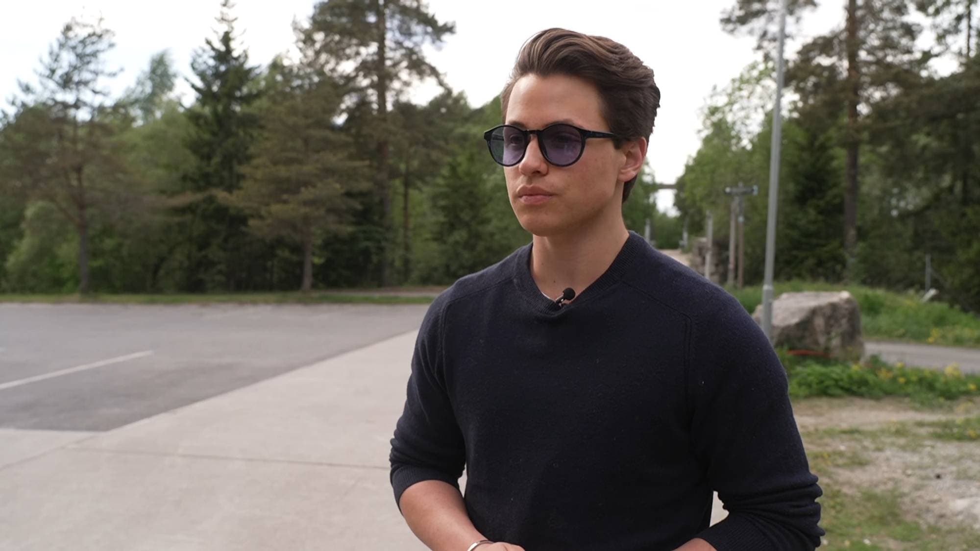 Ung mann i blå genser, står til høyre i bildet og ser mot venstre,. Han har kort brunt hår og blå solbriller på.