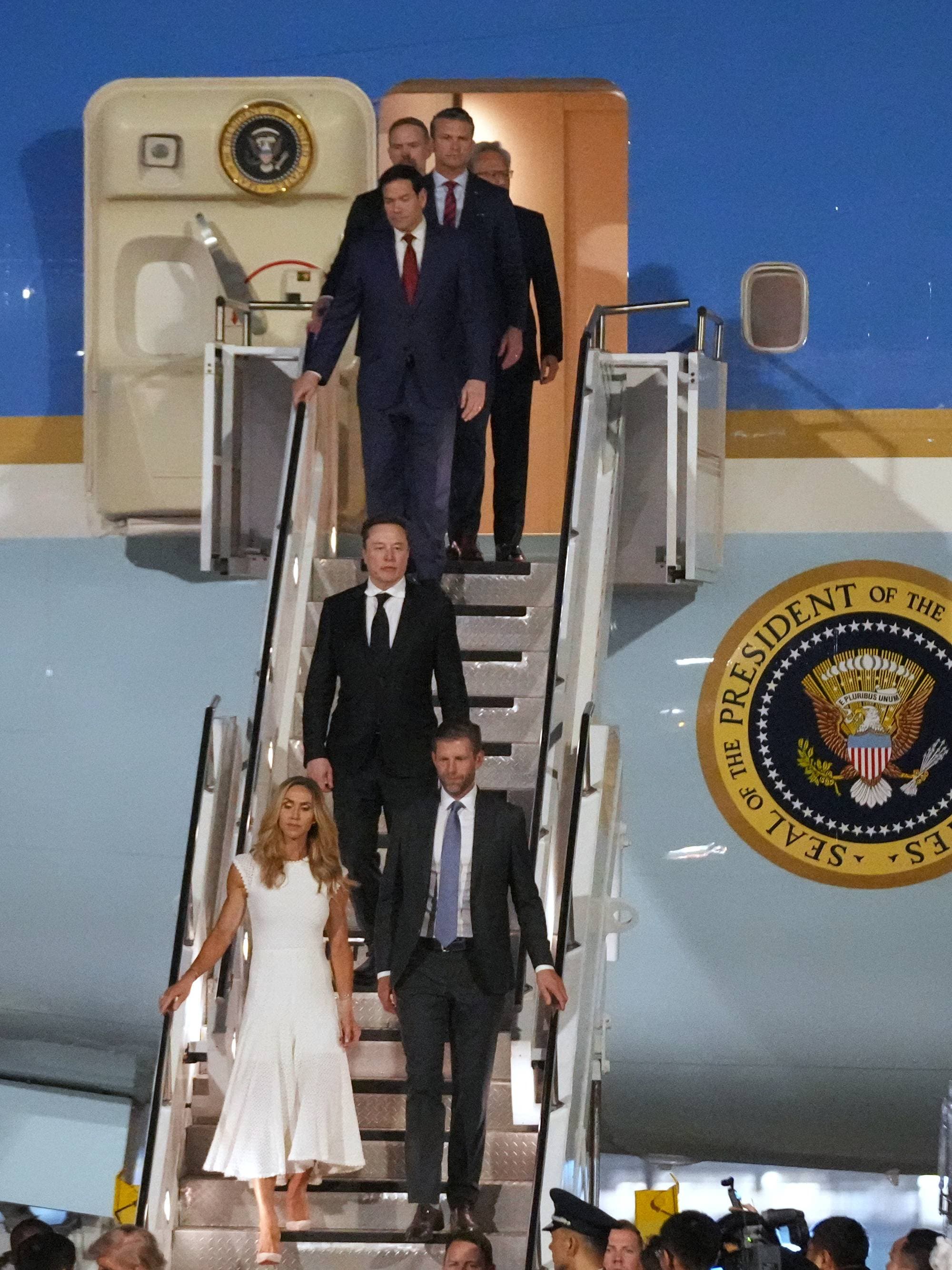 Elon Musk ankommer en flyplass med en kvinne i hvit kjole. De går ned en trapp fra et fly med presidentens segl. Flere personer, inkludert uniformerte, står på bakken. (Bildebeskrivelsen er laget av en KI-tjeneste)