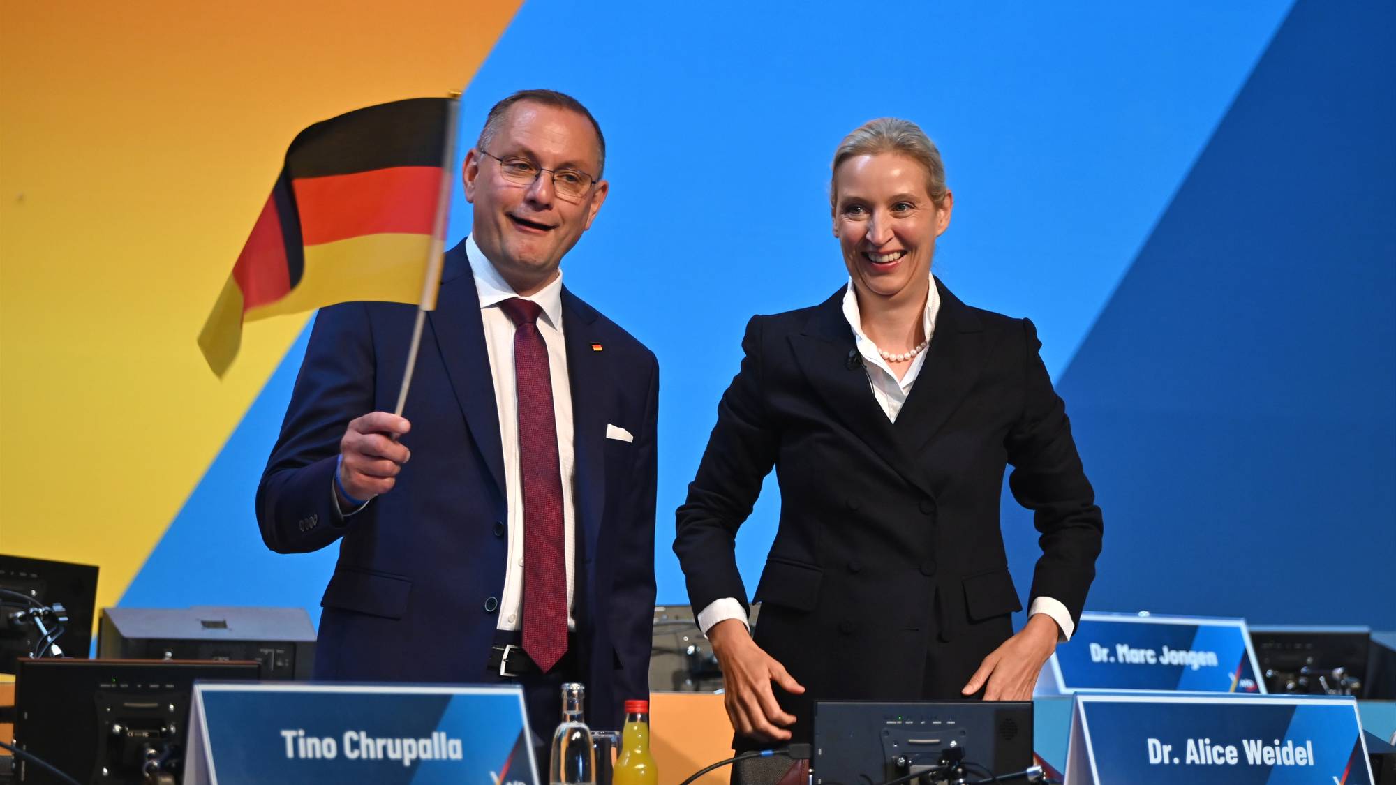 Tino Chrupalla og Alice Weidel fra AfD smiler foran en blå og gul bakgrunn. Chrupalla holder et flagg. (Bildebeskrivelsen er laget av en KI-tjeneste)