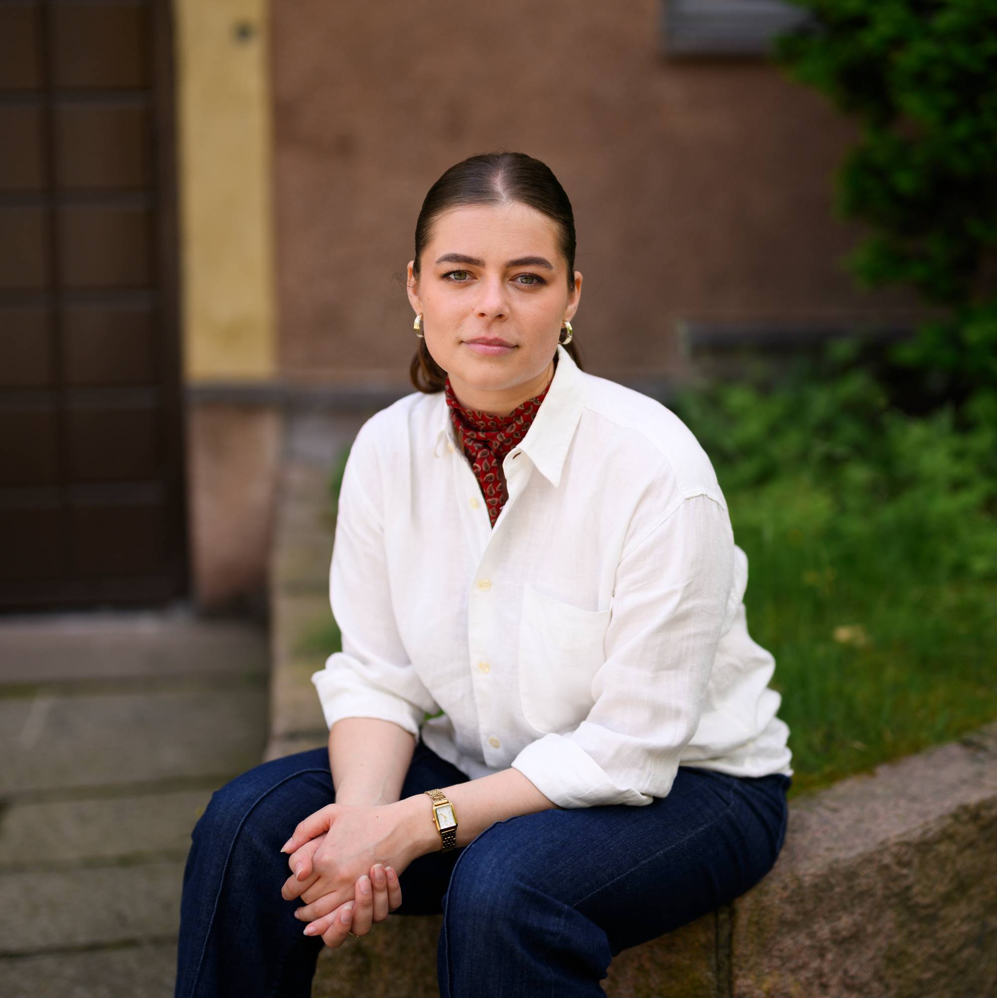 Kvinne med mørkt hår, hvit skjorte og mørke bukser sitter ute ved en murvegg. (Bildebeskrivelsen er laget av en KI-tjeneste)