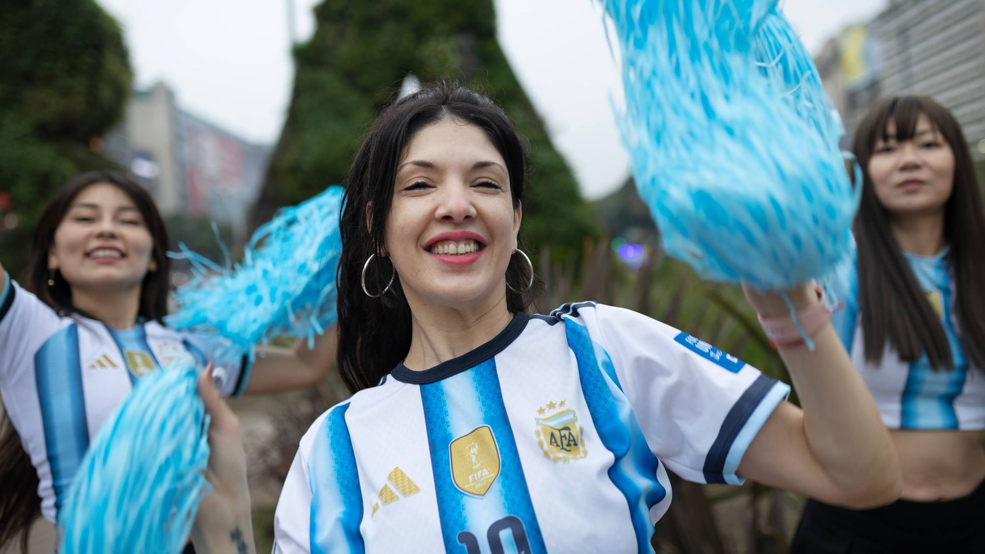 Tre kvinner i argentinske fotballdrakter vinker med lyseblå dusker i en bygate. 