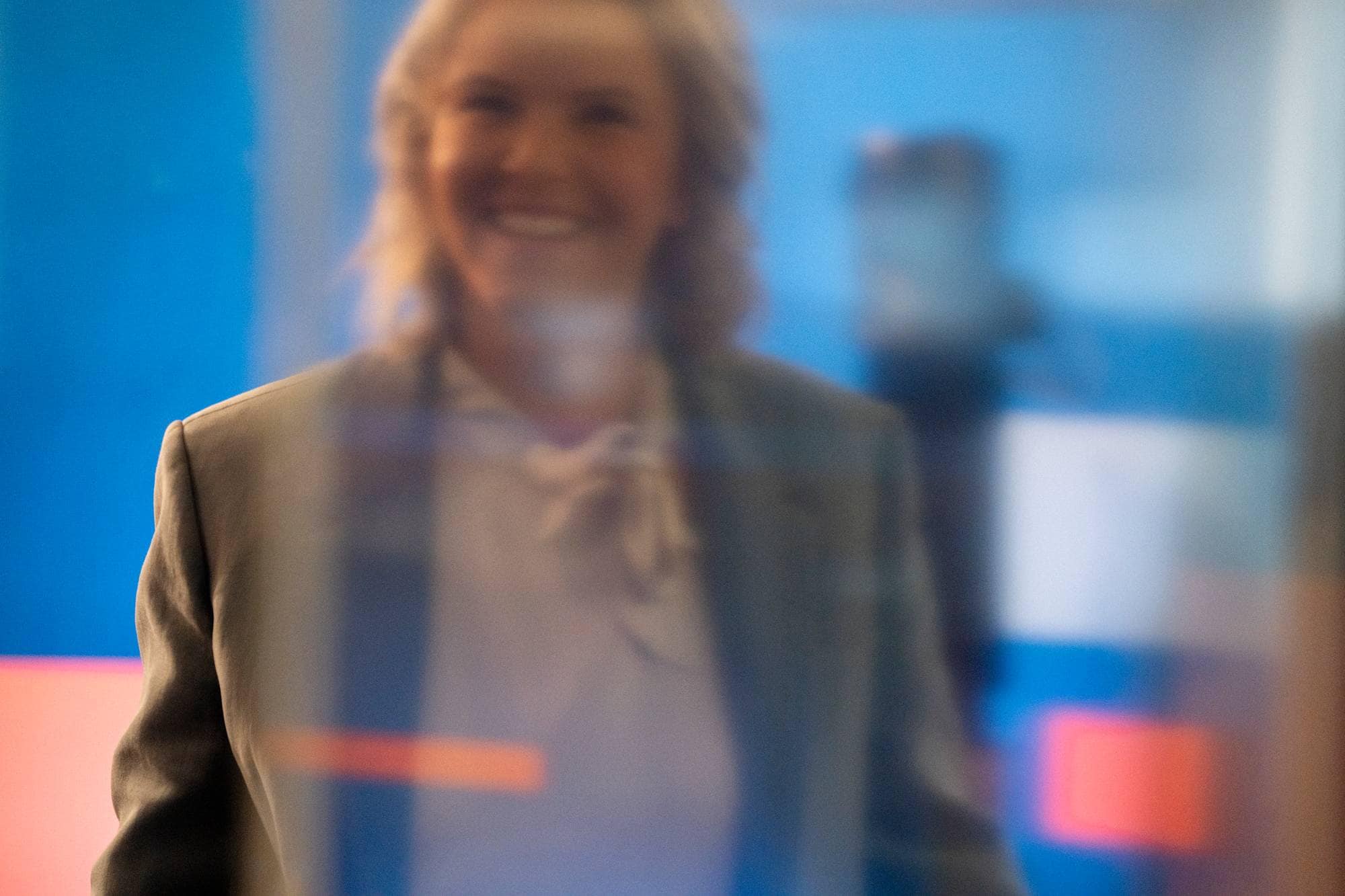 En kvinne smiler og ser mot kamera. Hun har på seg en blazer og en skjorte. Bakgrunnen er uskarp og har blå og oransje farger. Det er et uskarpt objekt til høyre for kvinnen. (Bildebeskrivelsen er laget av en KI-tjeneste)
