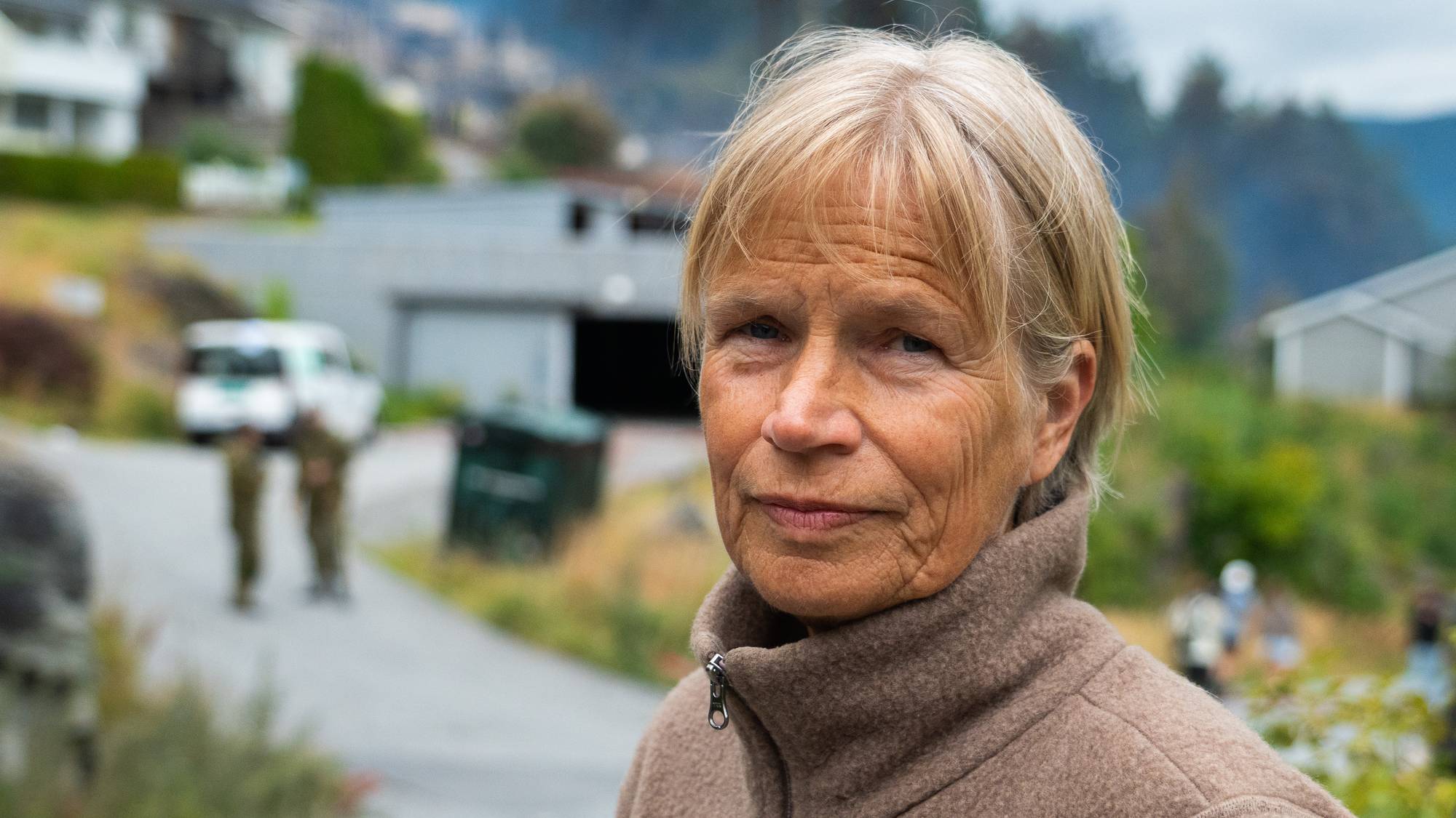 En kvinne med lyst, kort hår og beige fleece ser i kamera og står foran et boligfelt.