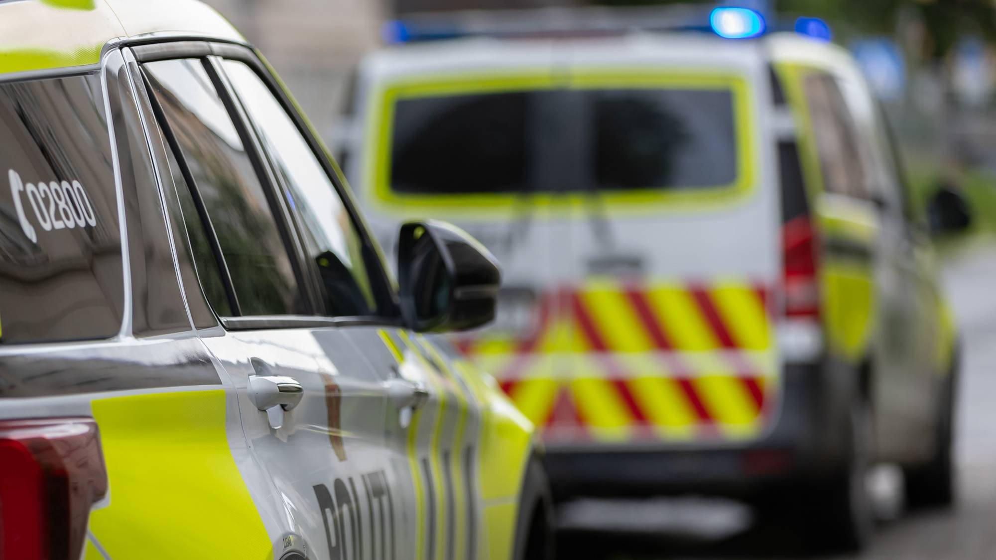 To politibiler står parkert. Vi ser bare deler av den bakerste, bilen men den er tydelig. Foran står en større politibil, men den har ikke fokus
