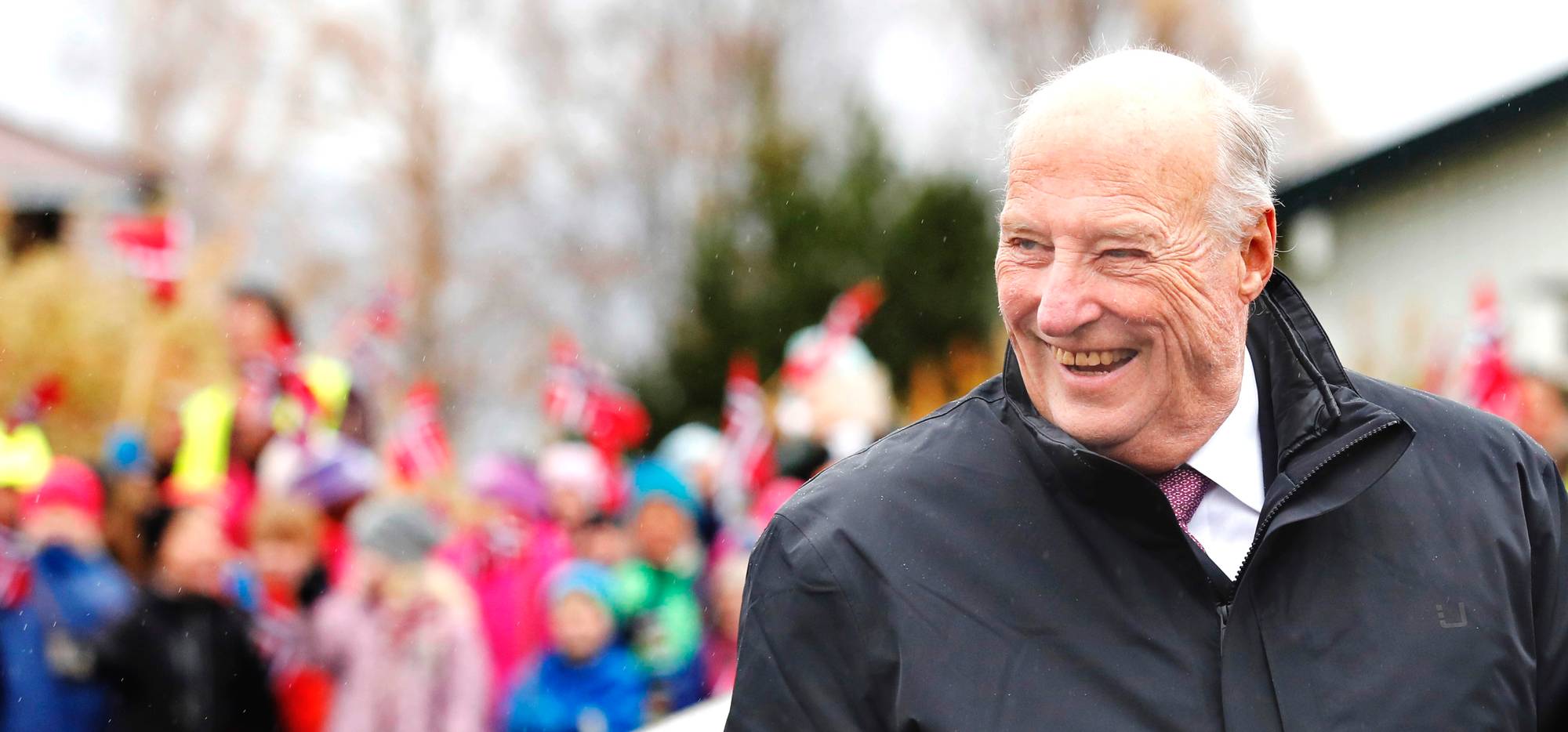 Kong Harald smiler, mens barn i bakgrunnen vifter med norske flagg