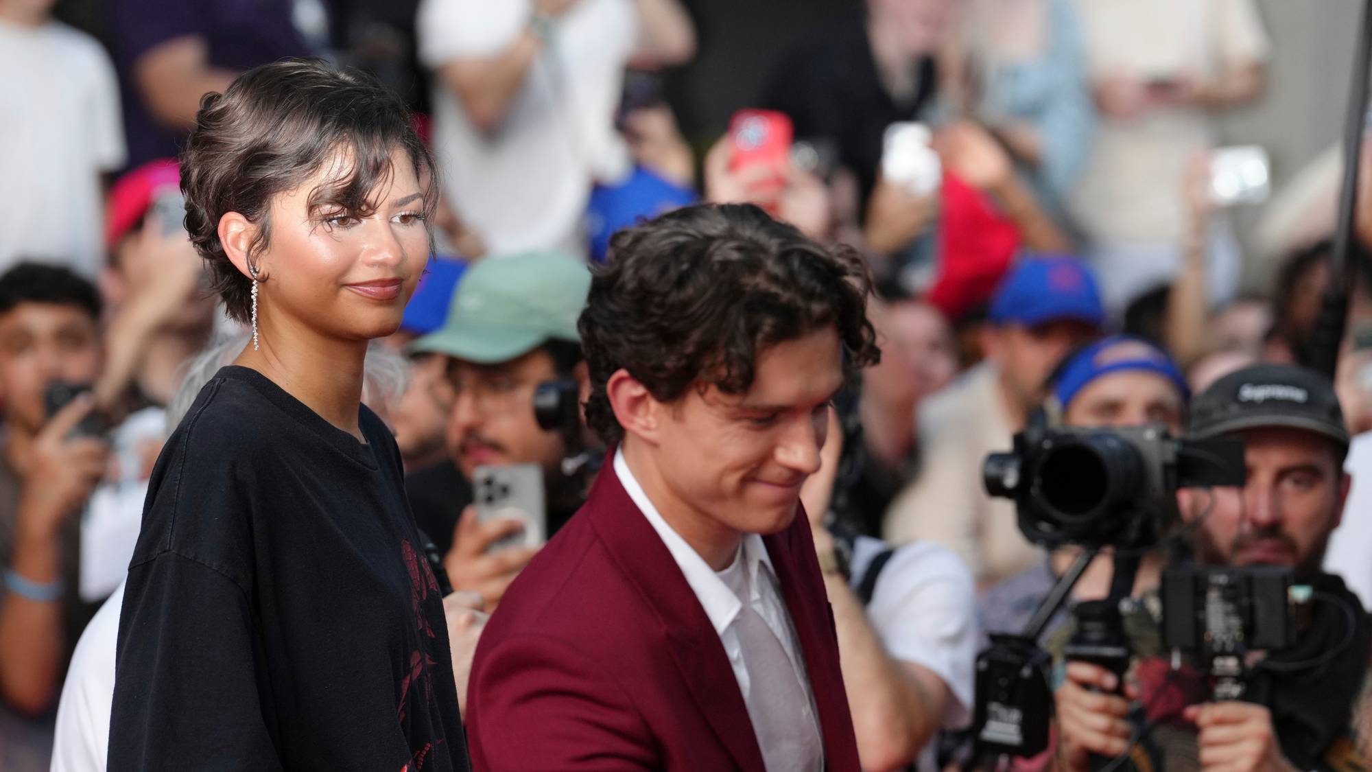 Mann med burgunderfarga dress hilser på publikum . Kvinne med svart kjole står bak og smiler.