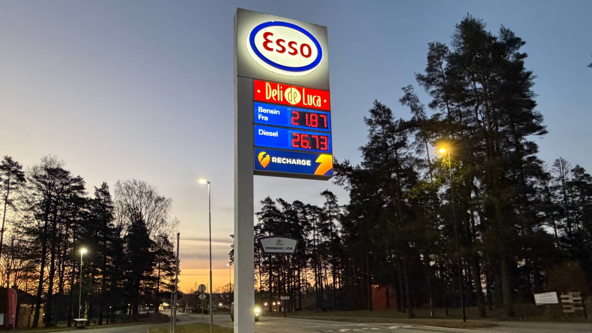 En Esso-stasjon med bensinpris på 21,87 kroner literen og diesel på 26,73 kroner literen. Ved siden av prisskiltet er det et skilt med "Minibank/ATM". Bildet er tatt i skumringen ved en vei omgitt av trær. 
