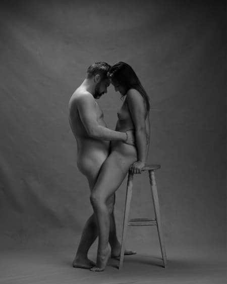 En naken mann står mellom bena på en naken kvinne som hviler rumpa på en høy krakk