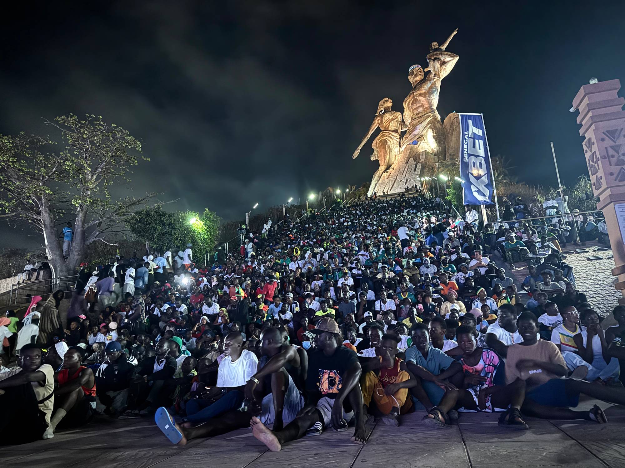 Stor folkemengde samlet foran monumentet afrikansk renessanse i Dakar, Senegal. Mange sitter på bakken og ser mot et podium, mens andre står eller sitter på tribuner.)