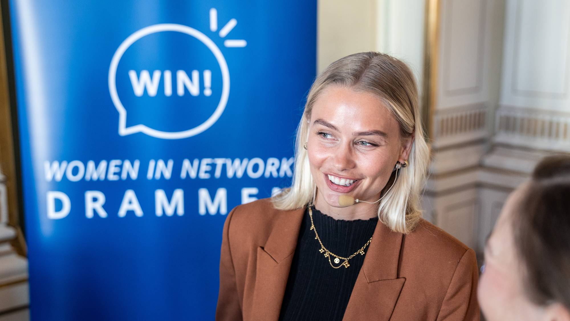 En kvinne med blondt hår smiler foran et blått banner med teksten "WIN! WOMEN IN NETWORK DRAMMEN". Hun har en brun blazer over en svart genser og et headset med mikrofon. I bakgrunnen sees en annen person og en dekorert vegg. (Bildebeskrivelsen er laget av en KI-tjeneste)