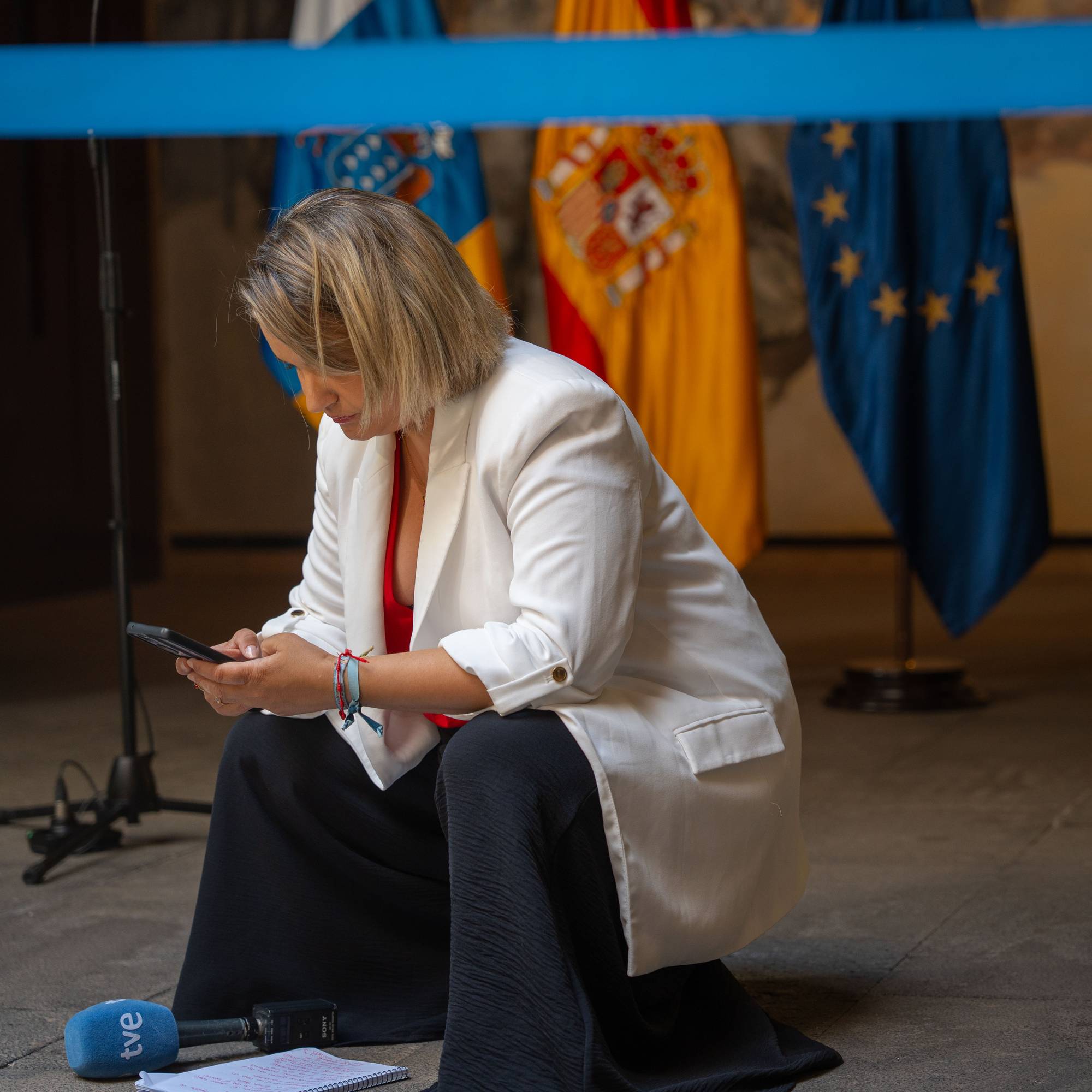 En kvinne sitter på huk og ser på telefonen. Hun har på seg en hvit blazer og en rød topp. Bak henne er det flere flagg, inkludert det spanske og EU-flagget. På bakken foran henne ligger en mikrofon med TVE-logo. (Bildebeskrivelsen er laget av en KI-tjeneste)
