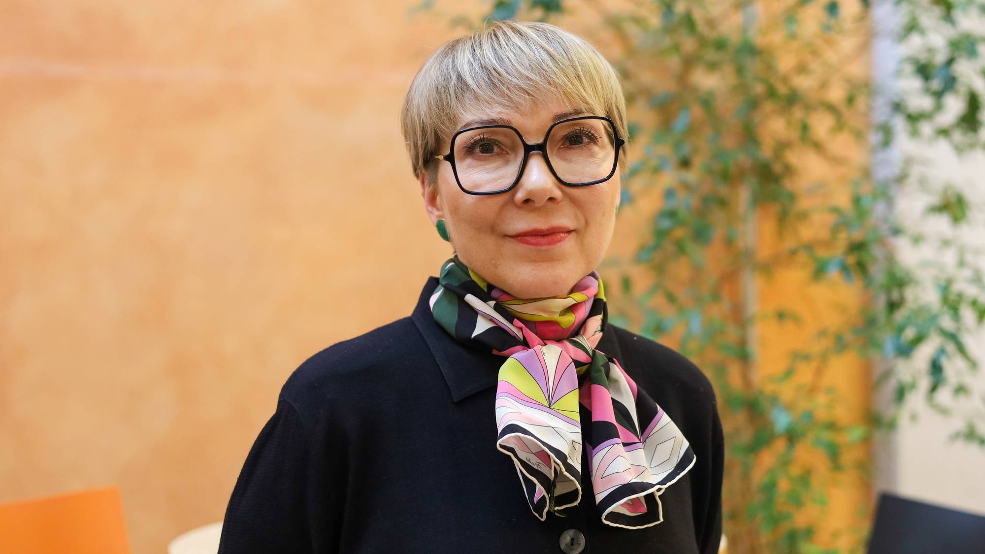 En kvinne med kort, blondt hår og store, svarte briller ser mot kamera. Hun har på seg et fargerikt skjerf og en mørk jakke. Bak henne er en oransje vegg og grønne planter. (Bildebeskrivelsen er laget av en KI-tjeneste)