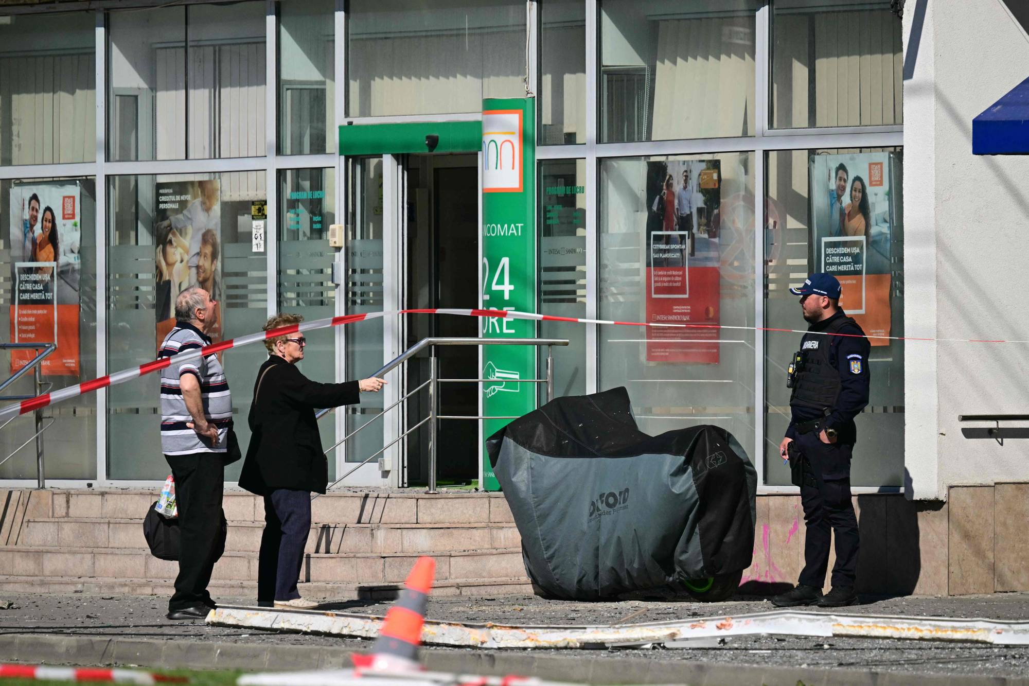 En politibetjent står utenfor en bank med avsperringstape. To sivile ser mot banken. En motorsykkel er tildekket av et grått trekk. Utenfor banken ligger rester av glass og annet materiale på bakken. (Bildebeskrivelsen er laget av en KI-tjeneste)