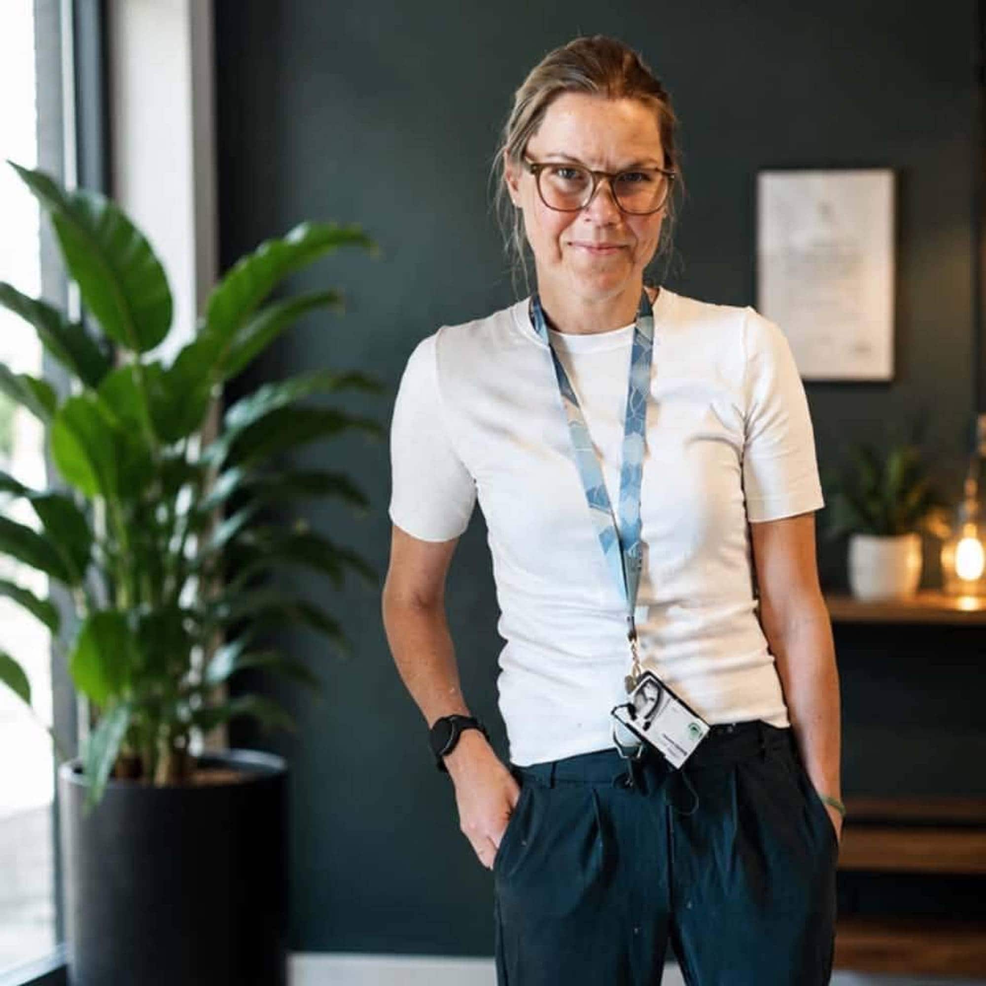 Kvinne med briller, hvit t-skjorte og adgangskort rundt halsen står innendørs ved en grønn plante. (Bildebeskrivelsen er laget av en KI-tjeneste)